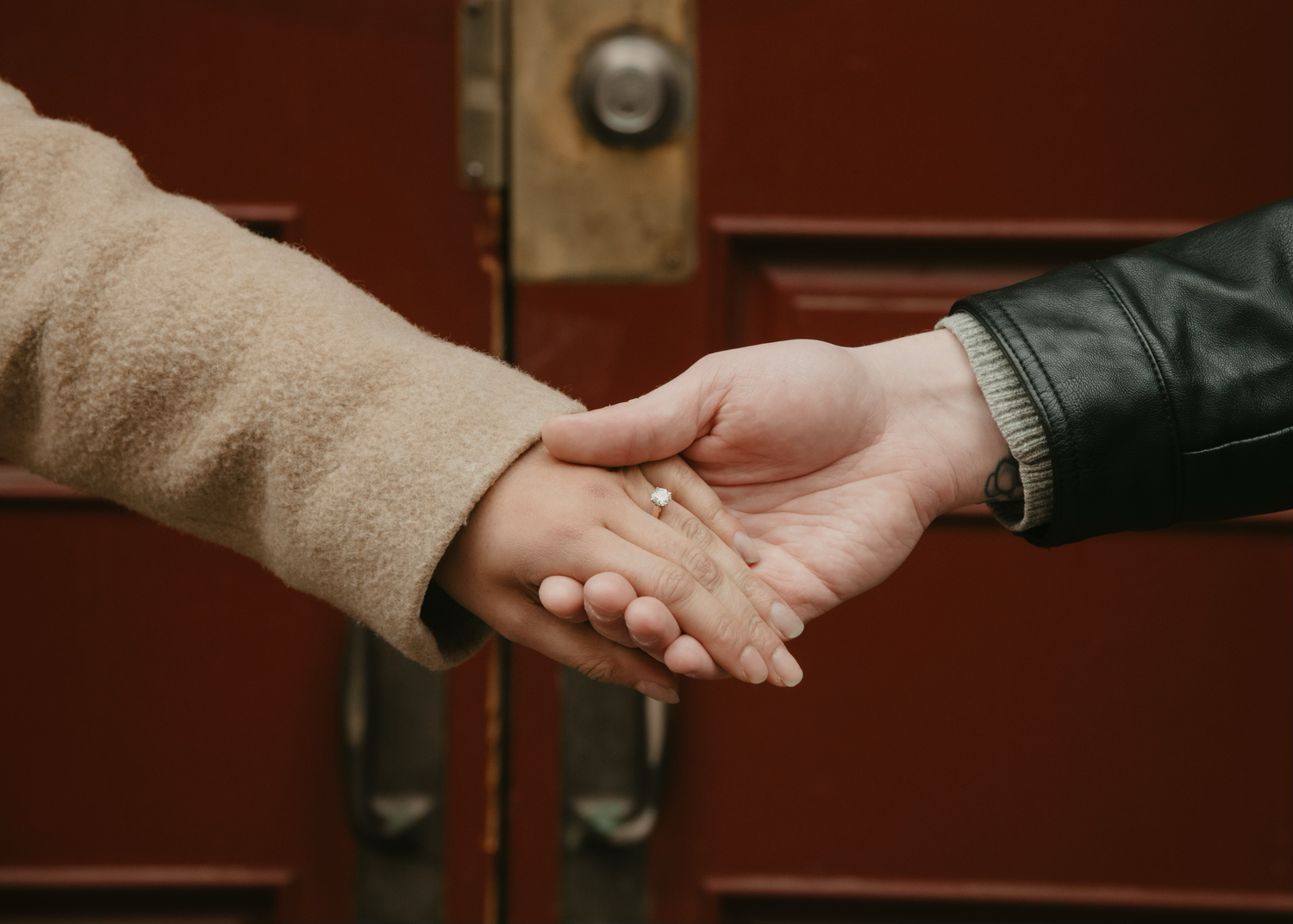 Two hands joining together against a red door background in an intimate gesture. Calgary Alberta Engagement Photographer.