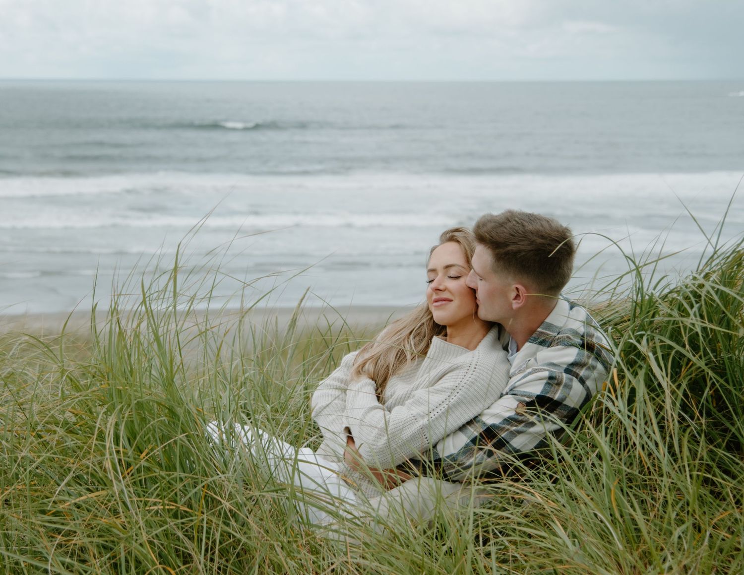 Couple cuddling on grassy oceanside cliff while enjoying the sea view on Oregon Coast. Calgary Lifestyle and Engagement Photographer.