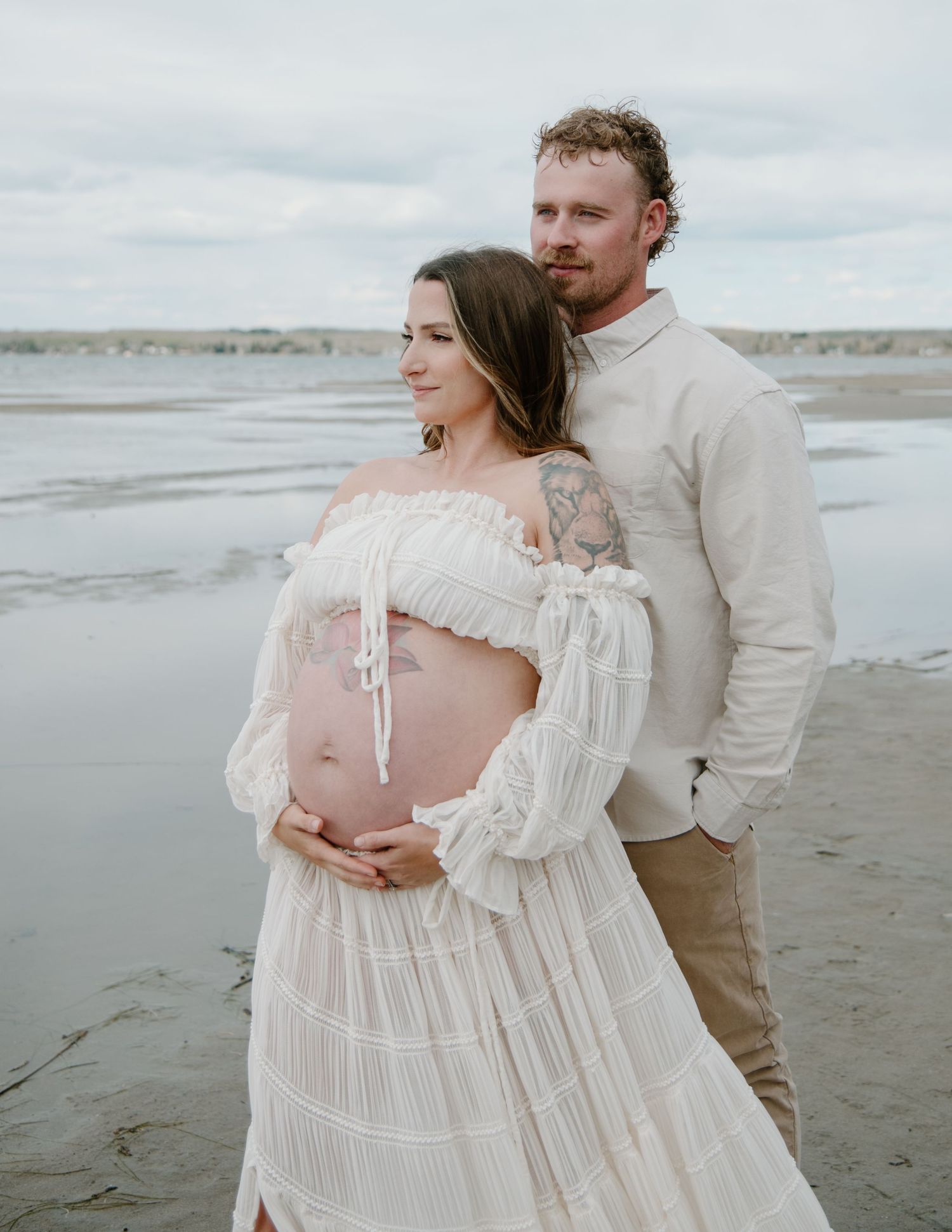 A maternity beach portrait featuring a couple in coordinated neutral outfits against an overcast sky. Calgary Alberta Maternity Photographer