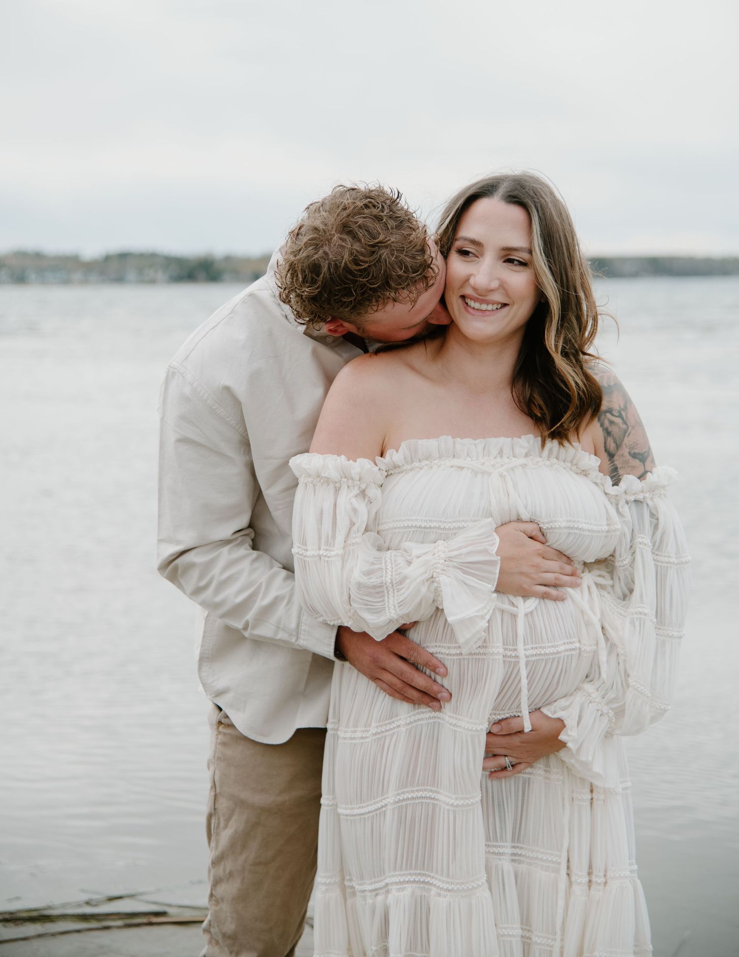 A tender moment between a couple in light-colored clothing standing by the ocean. Calgary Alberta Maternity Photographer.