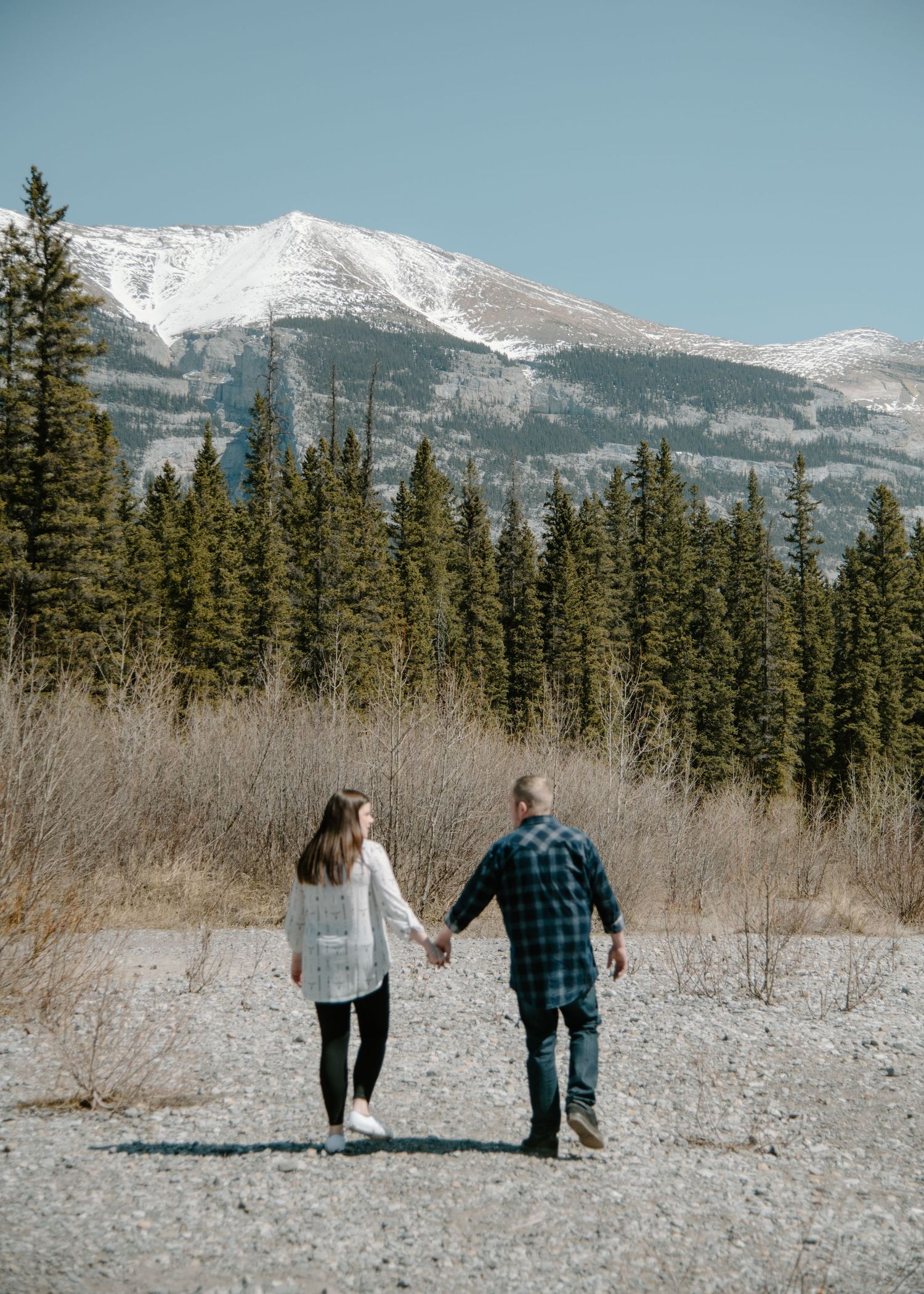 A couple walking hand in hand through a snowy mountain landscape with evergreen trees. Calgary Alberta Engagement Photographer.
