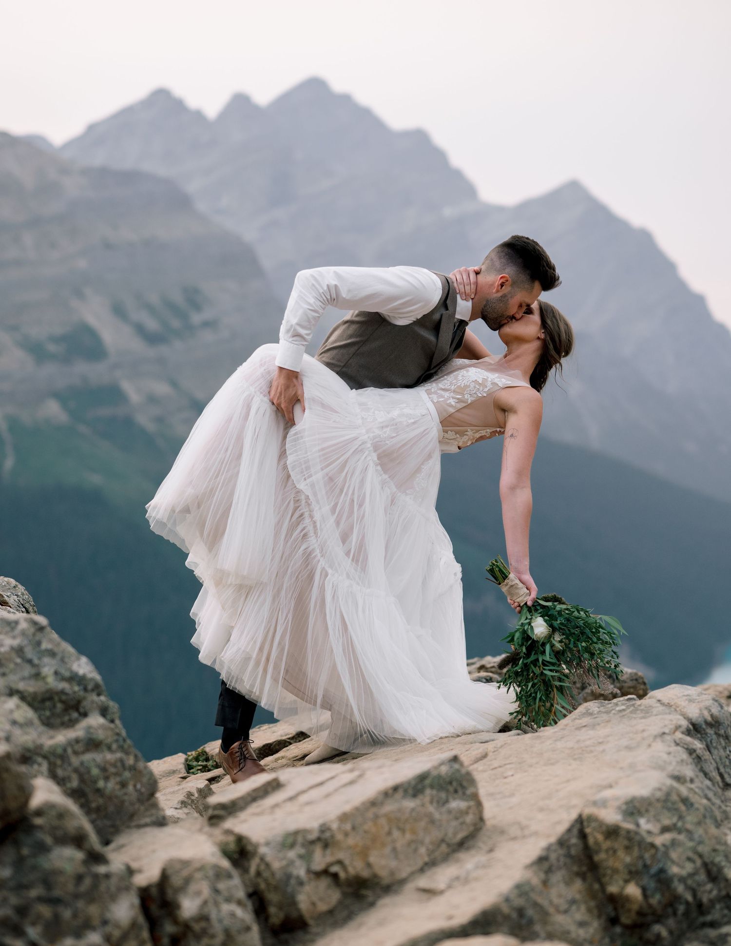 A romantic wedding couple embrace on a mountain ledge with majestic rocky peaks in the background.