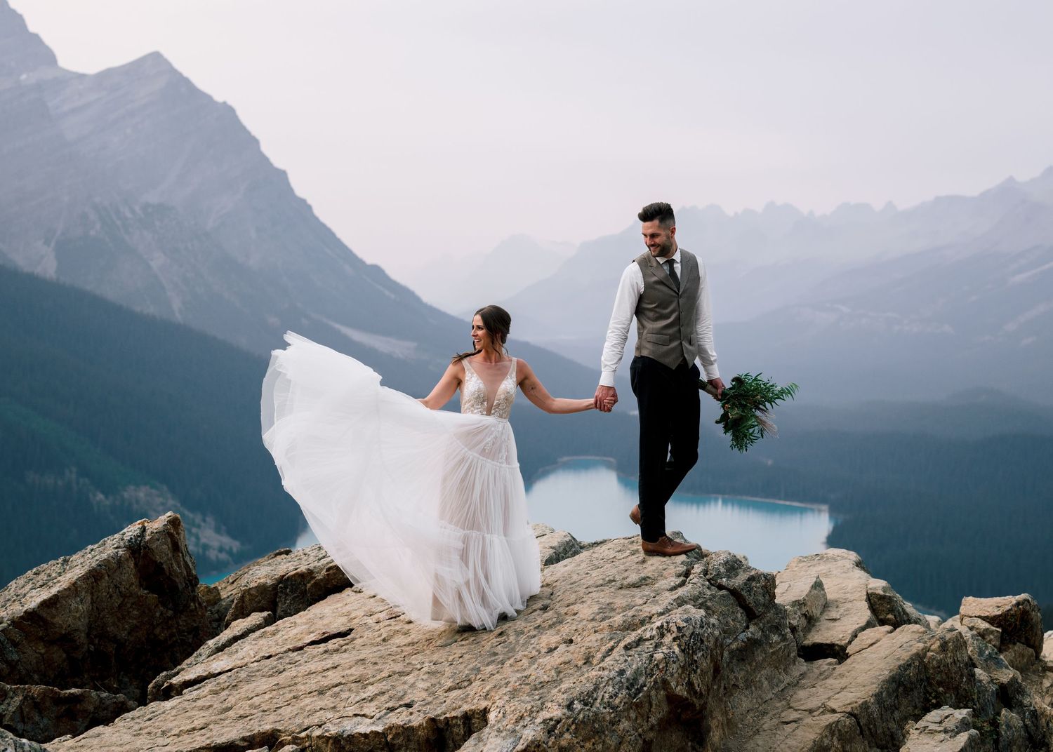 A couple poses on a mountain peak overlooking a lake and mountain range during their wedding photoshoot.