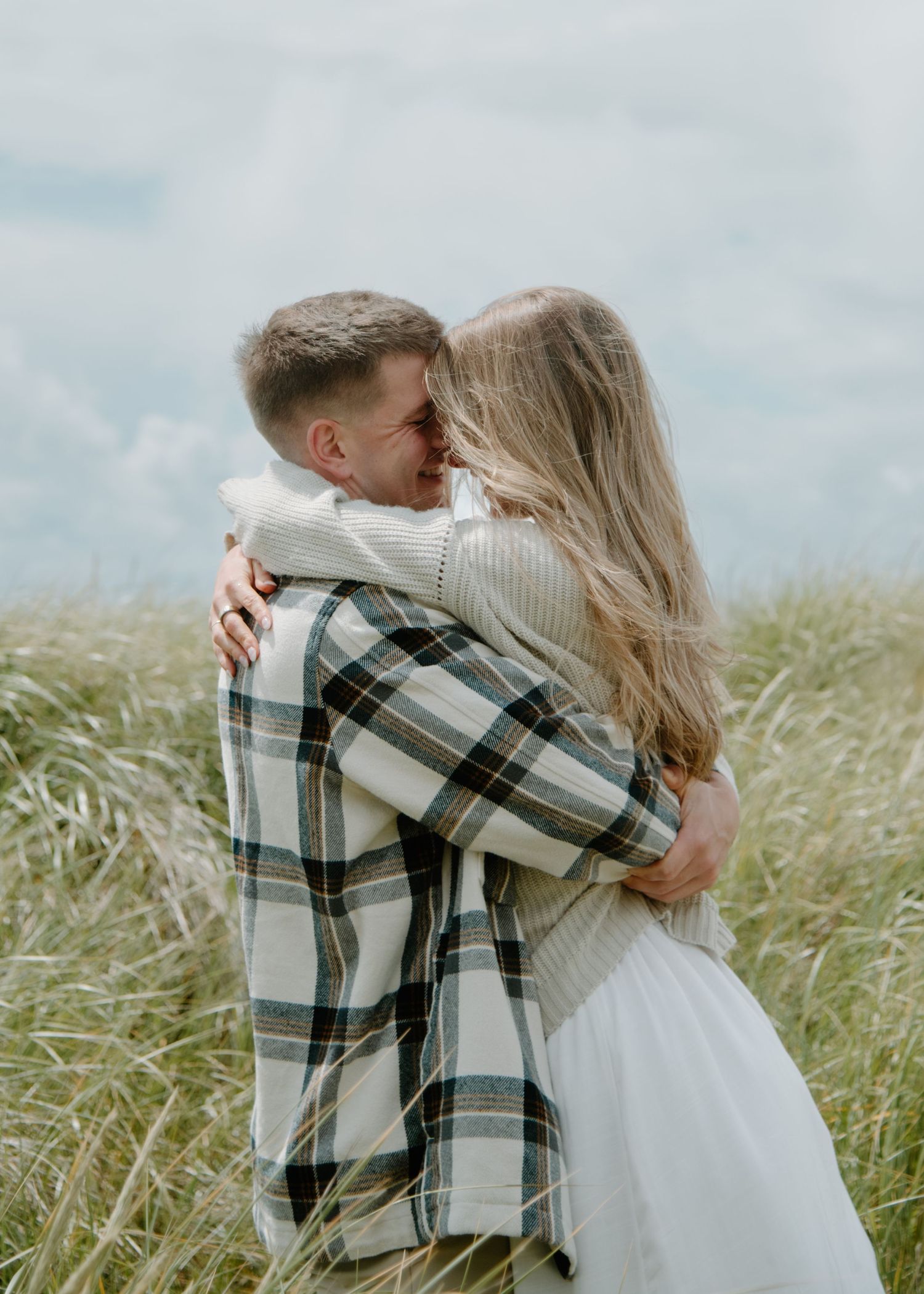 An intimate embrace on a windswept coastal hillside with flowing beach grass on Oregon Coast. Calgary Lifestyle and Engagement Photographer.