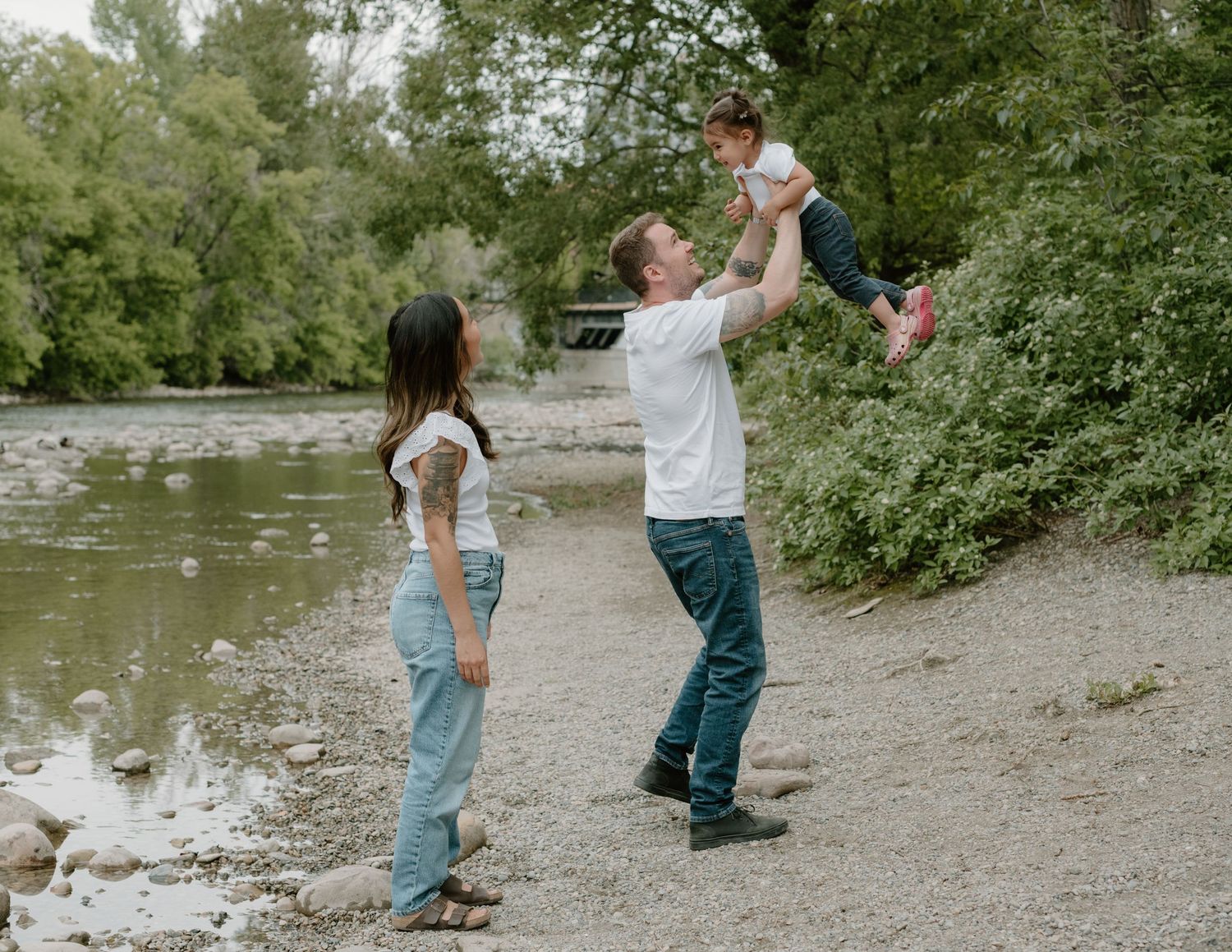 Family playing together on a rocky riverbank surrounded by green foliage at Lindsey Park, Calgary. Calgary Maternity Photographer.