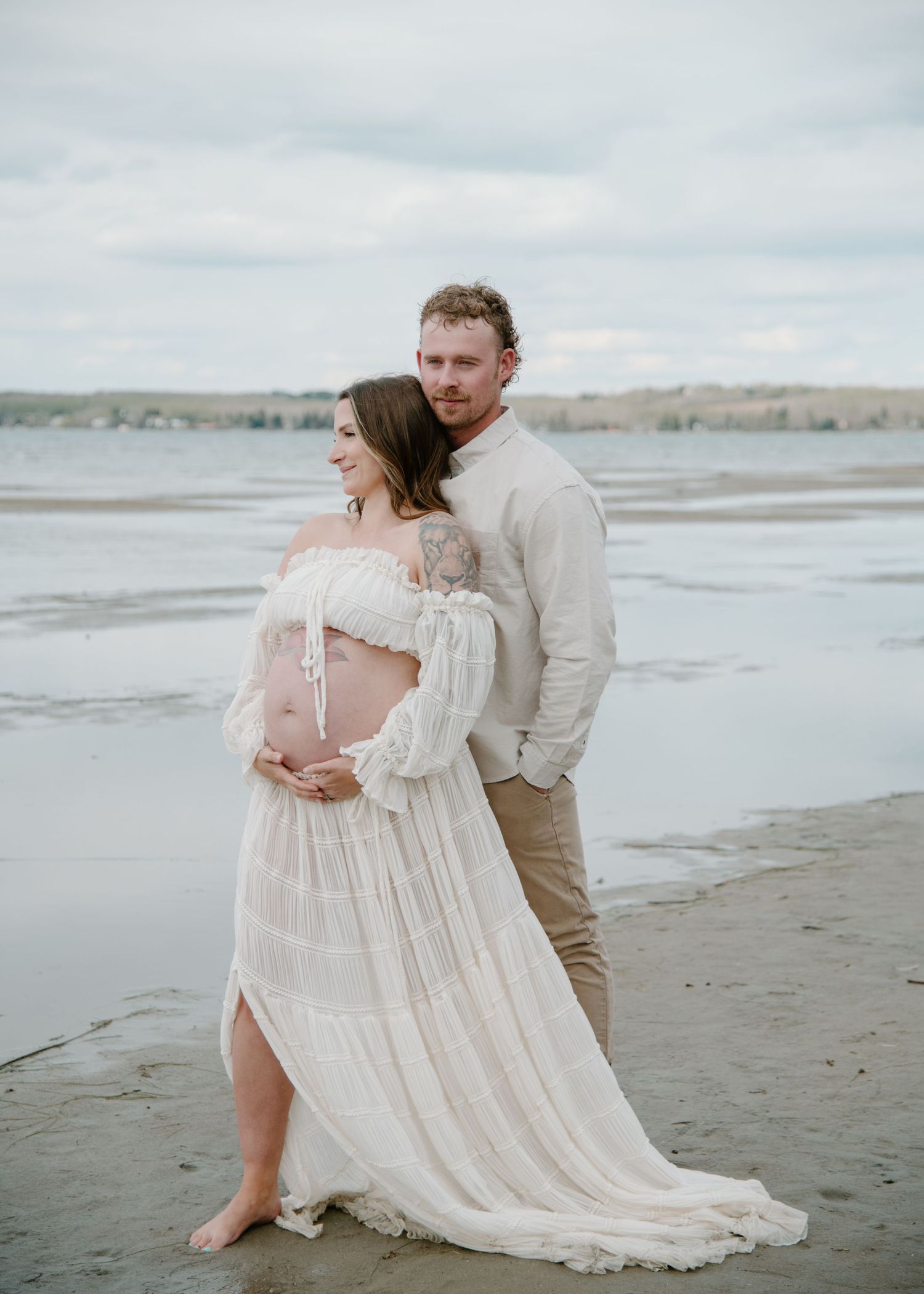 Couple embraces during a beachside maternity session wearing coordinated neutral tones. Calgary Alberta Maternity Photographer.