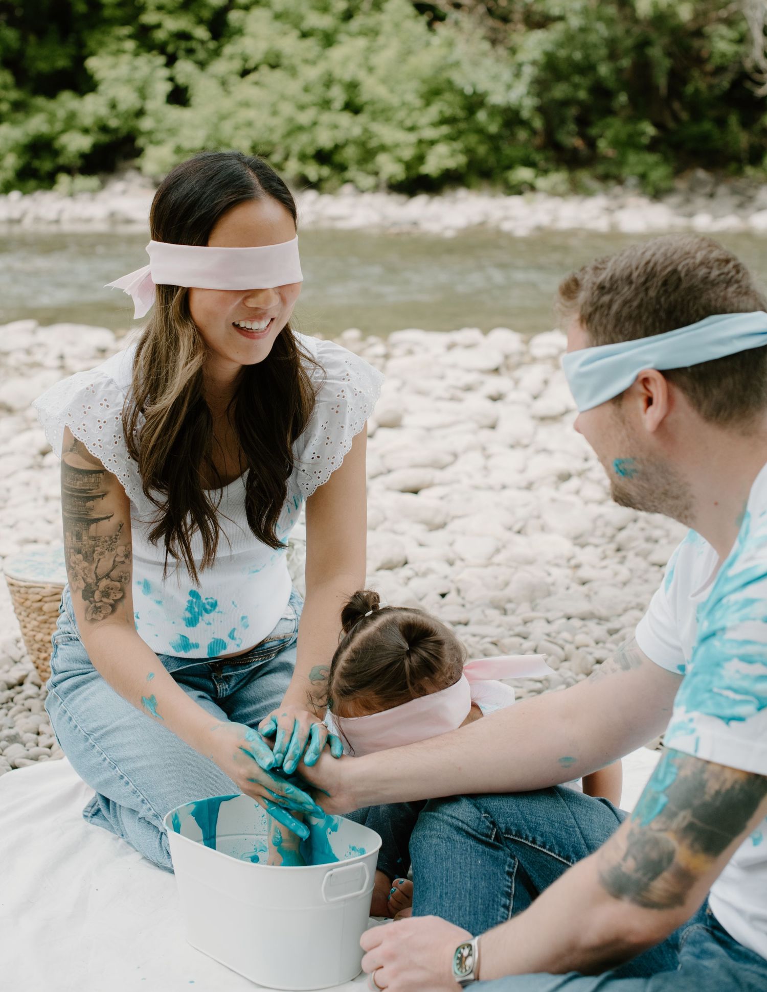 A family wearing blindfolds and paint-stained clothes plays a messy game by the river. Calgary Alberta Family and Maternity Photographer.