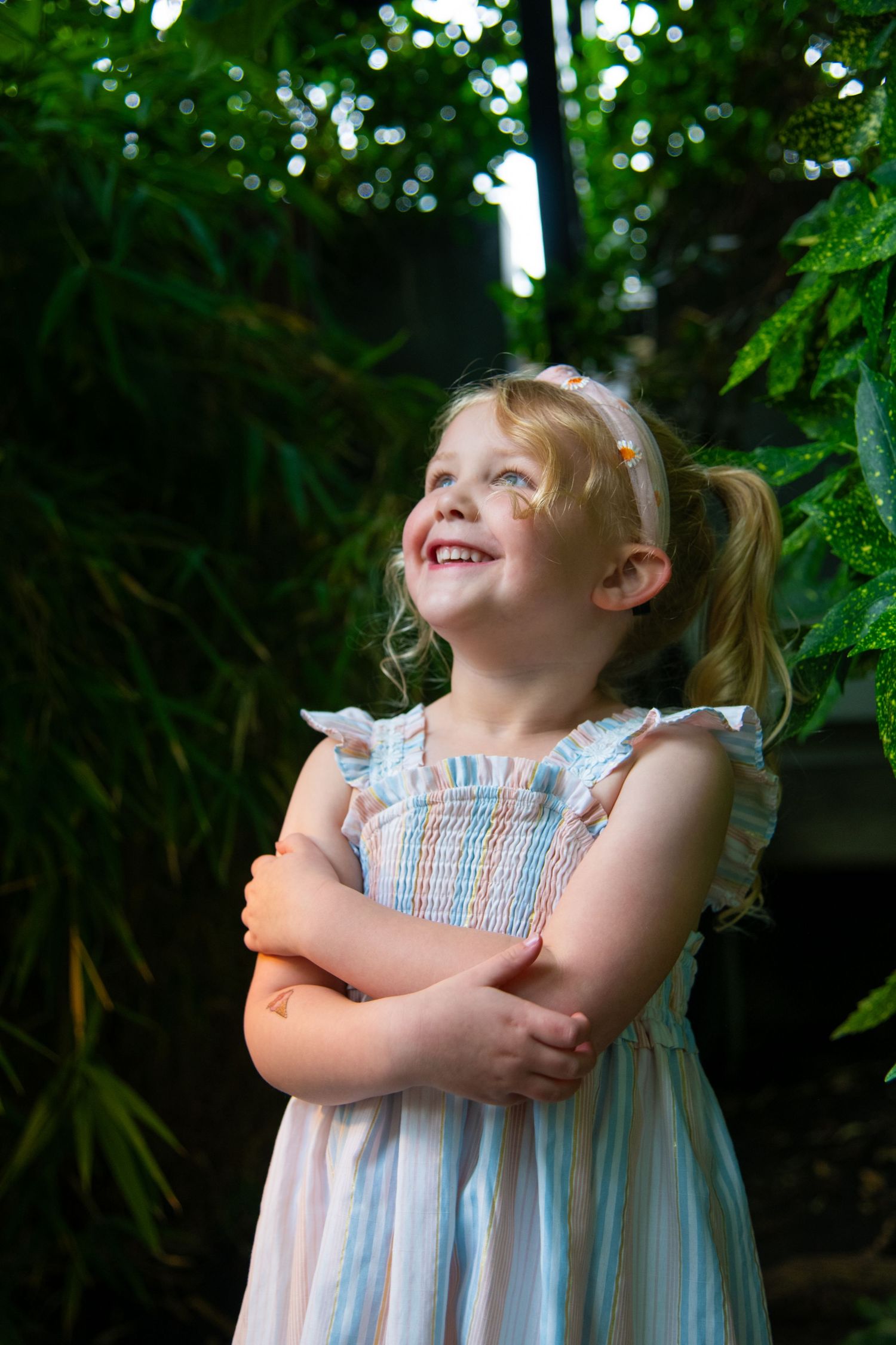 Young child in blue and white striped dress smiles joyfully against lush green foliage background. Calgary Alberta Family Photographer.