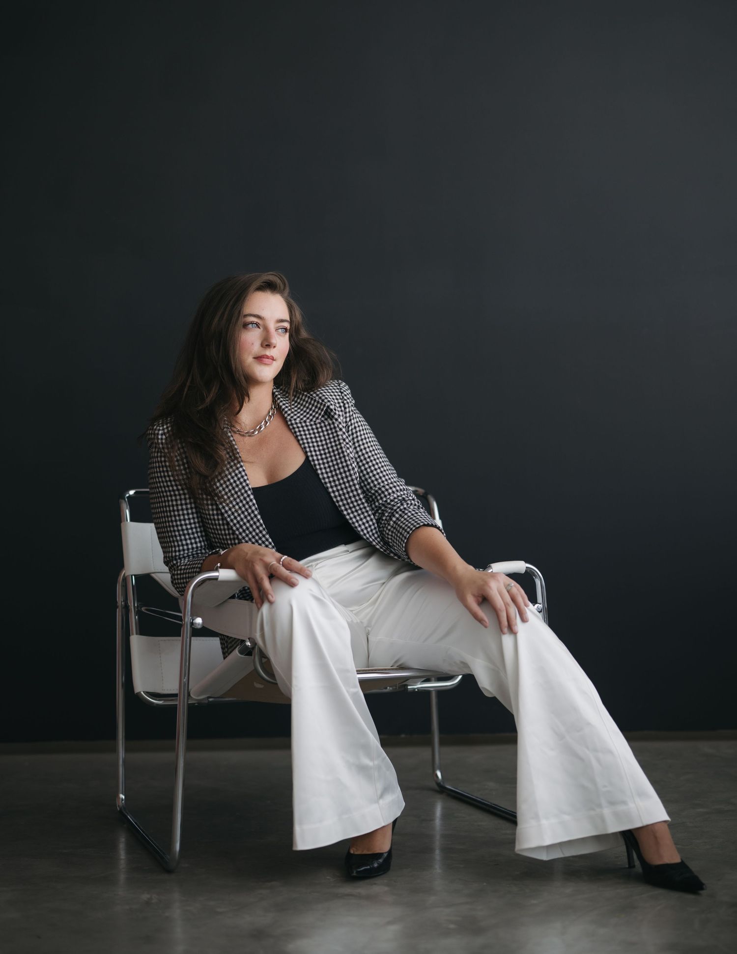 Professional studio portrait of someone seated in a modern chair wearing a patterned blazer and white pants against dark background.