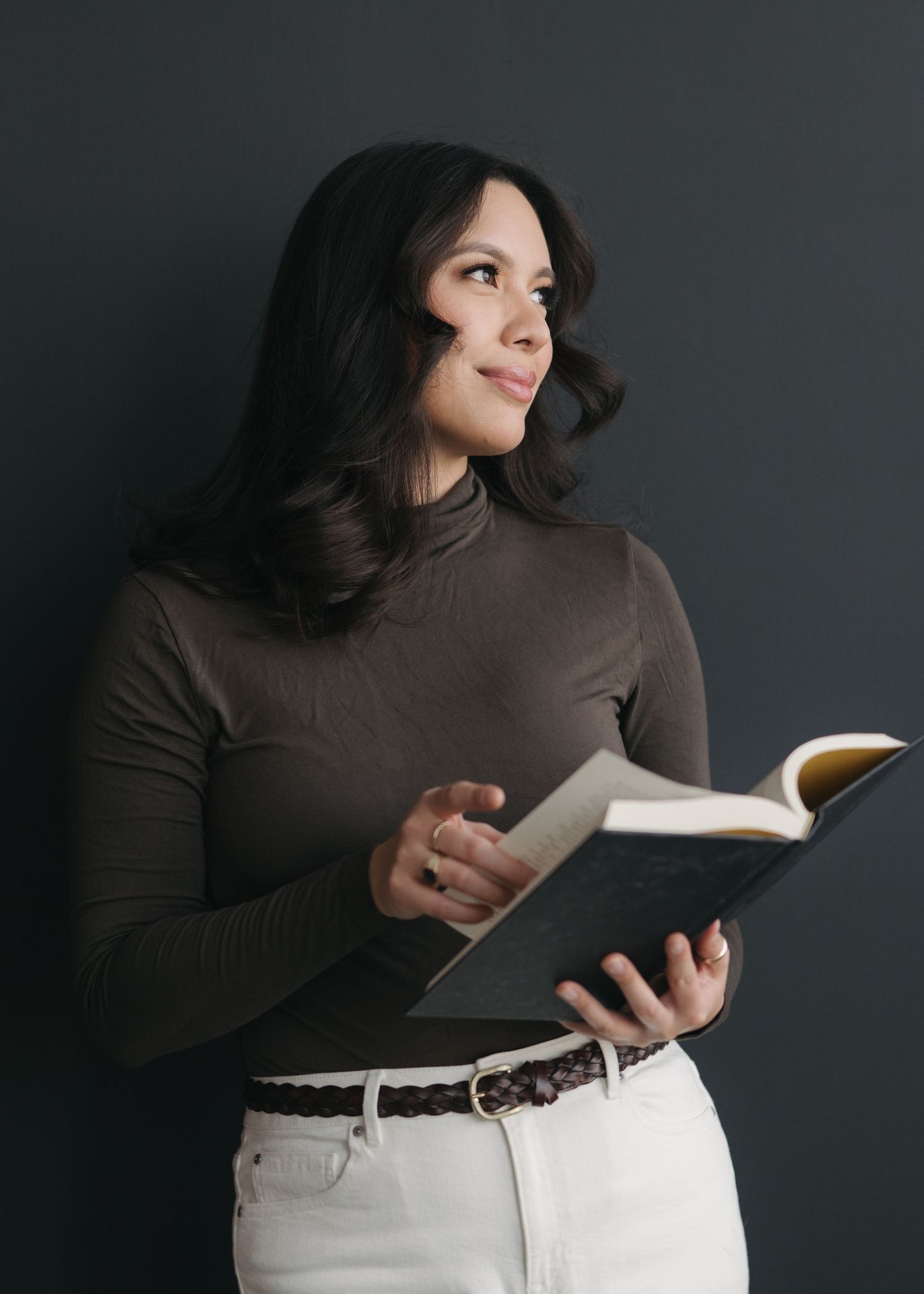 Person in brown turtleneck and white pants reads book against dark background in artistic portrait series.