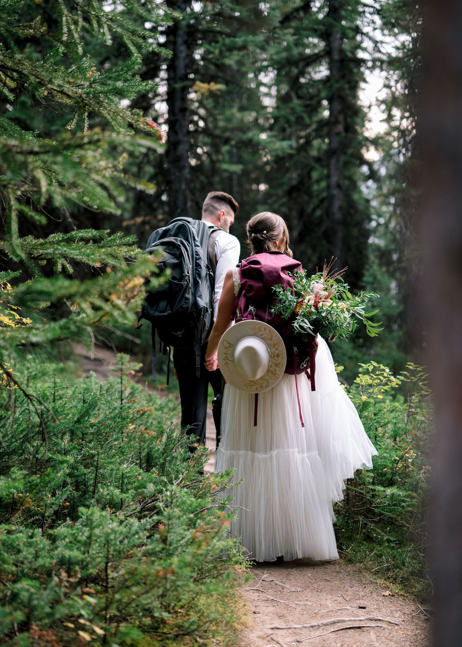 A couple in wedding attire walking together on a forest path surrounded by lush evergreen trees.