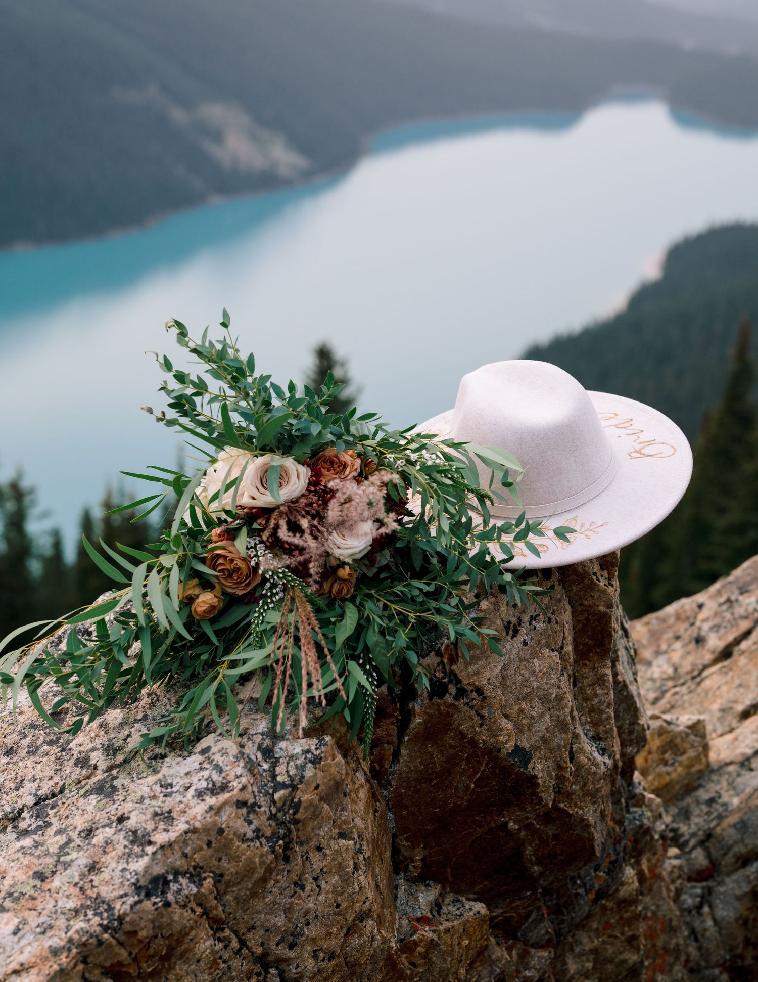 A rustic wedding bouquet with greenery and a white cowboy hat perched on rocks overlooking a turquoise alpine lake.