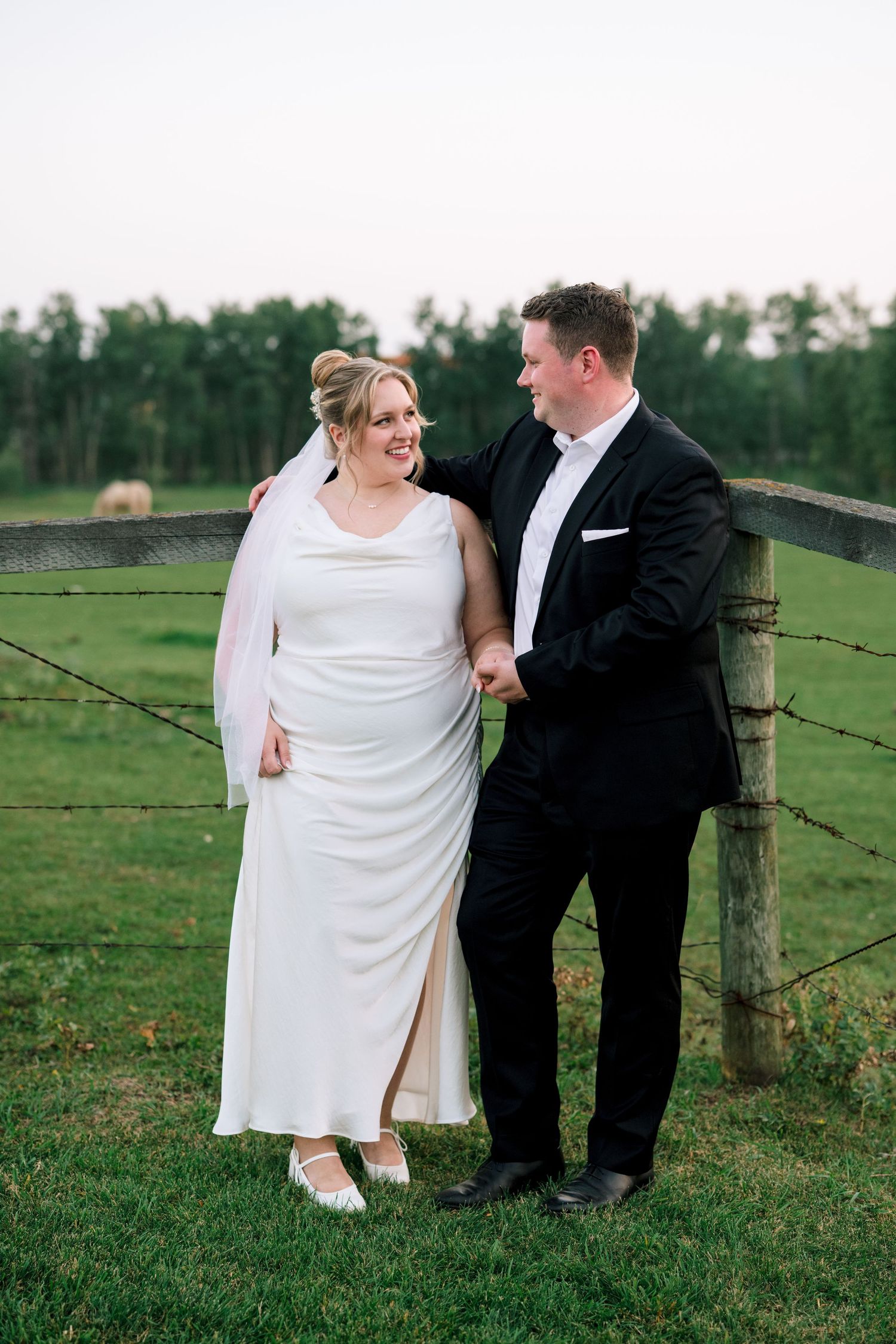 Wedding couple shares intimate moment against rustic farm fence on their special day in lush green field.