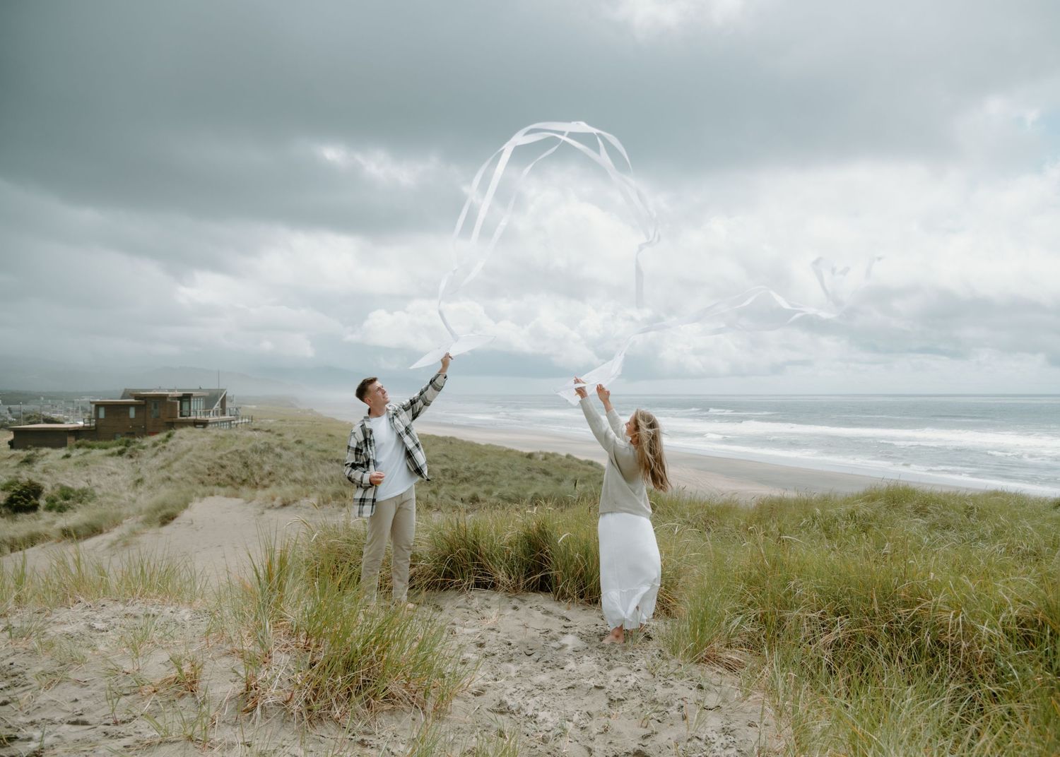 Two people throw flowing white fabric into the air against a moody coastal backdrop on Oregon Coast. Calgary Lifestyle and Engagement.