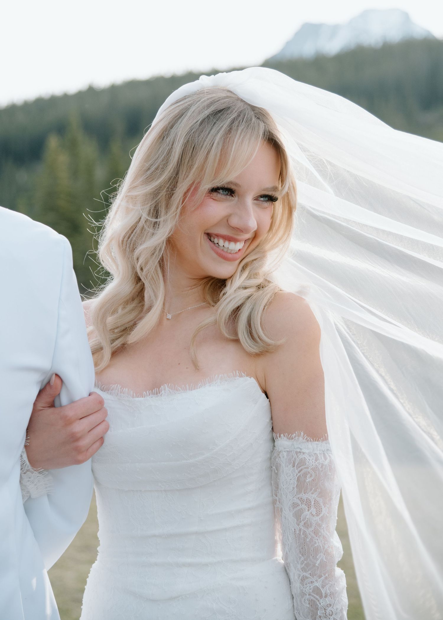Bride in white dress and veil smiles radiantly against mountain backdrop. Calgary Wedding Photographer