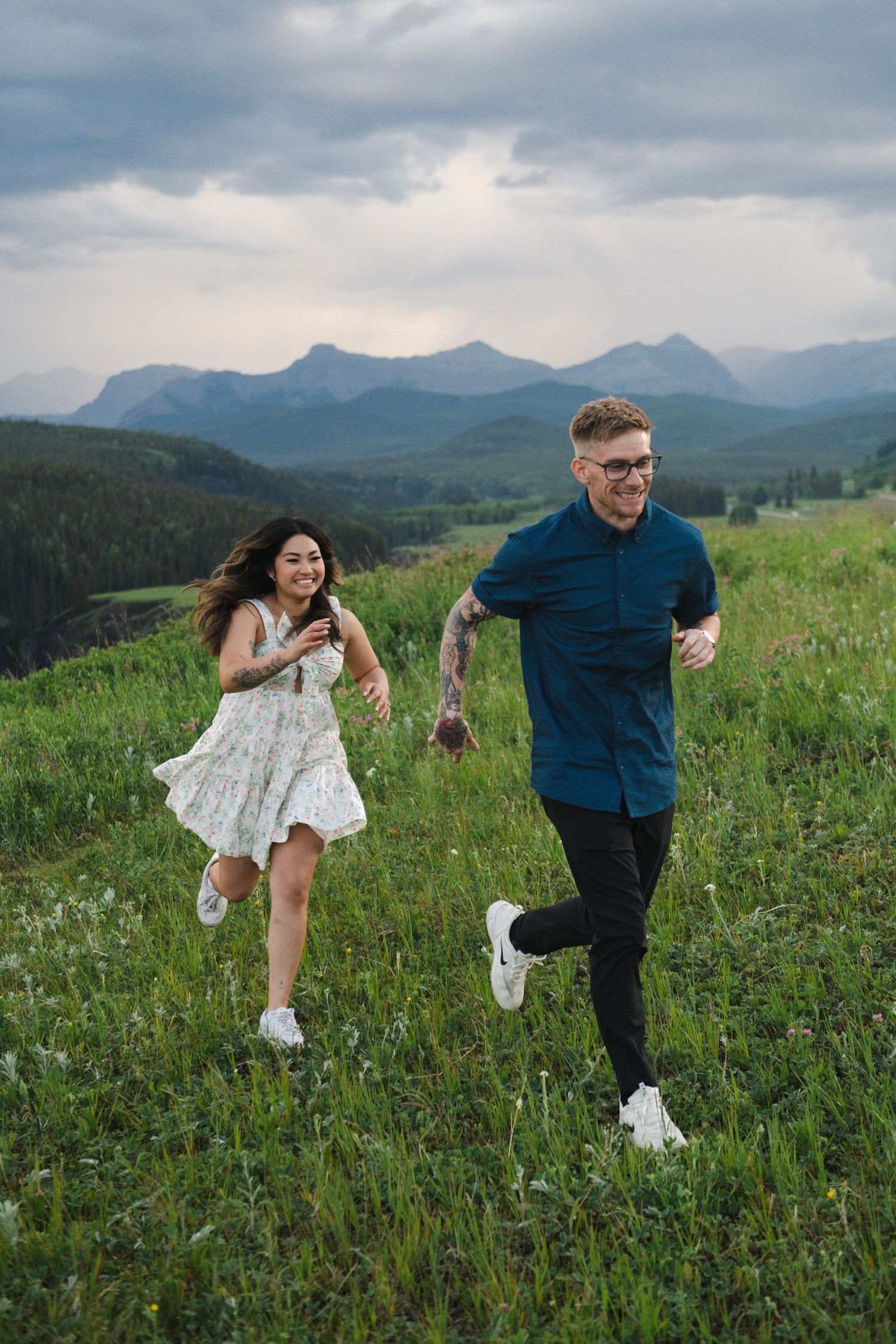 Two people run playfully through a mountain meadow with dramatic cloudy skies in Calgary. Calgary Lifestyle and Engagement Photographer.