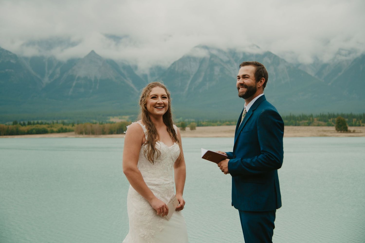 Joyful wedding moment captured against dramatic mountain landscape and calm waters. Calgary Wedding Photographer