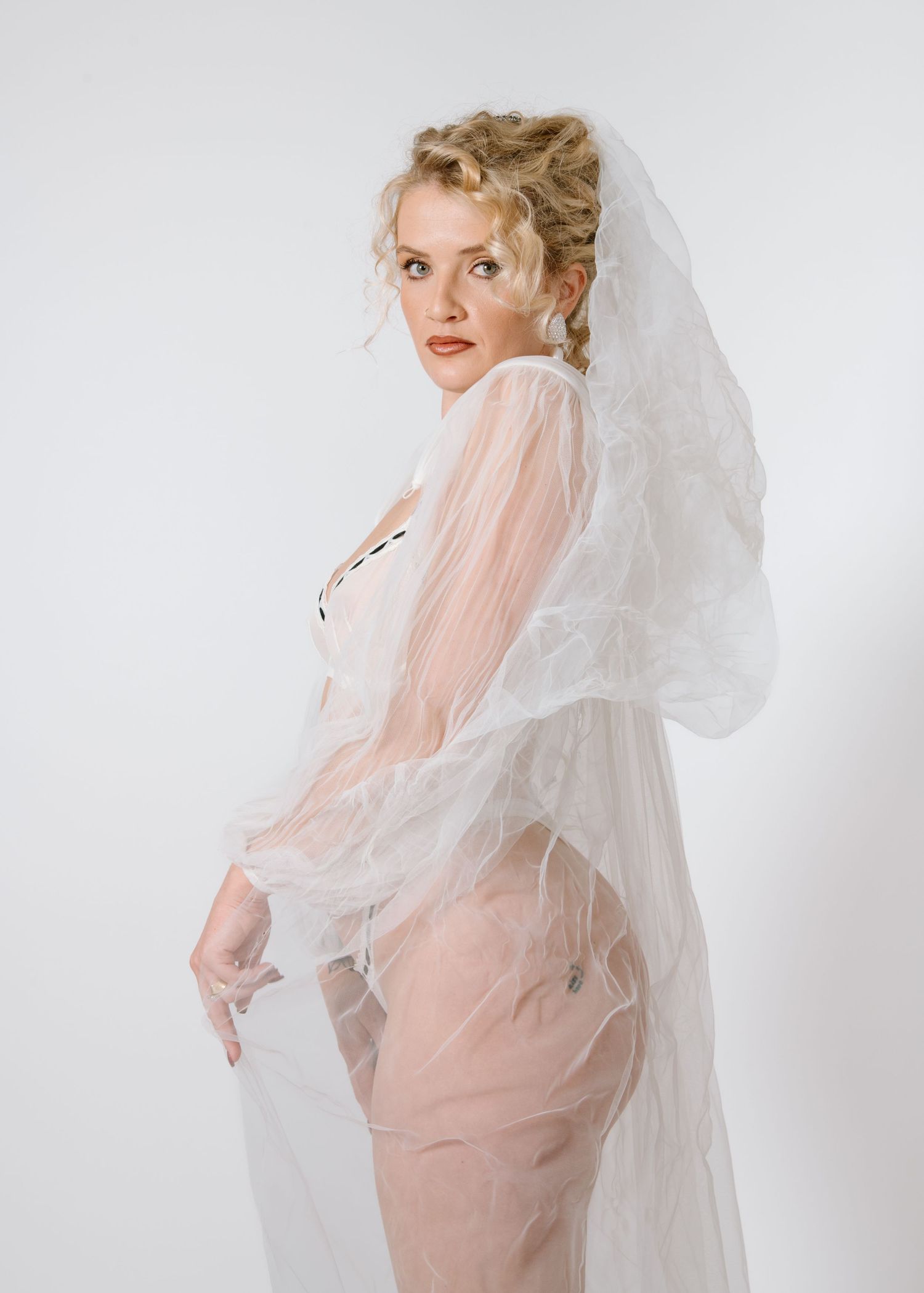A bride in a sheer veil poses against a white backdrop in romantic boudoir style. Calgary Bridal Boudoir Photographer