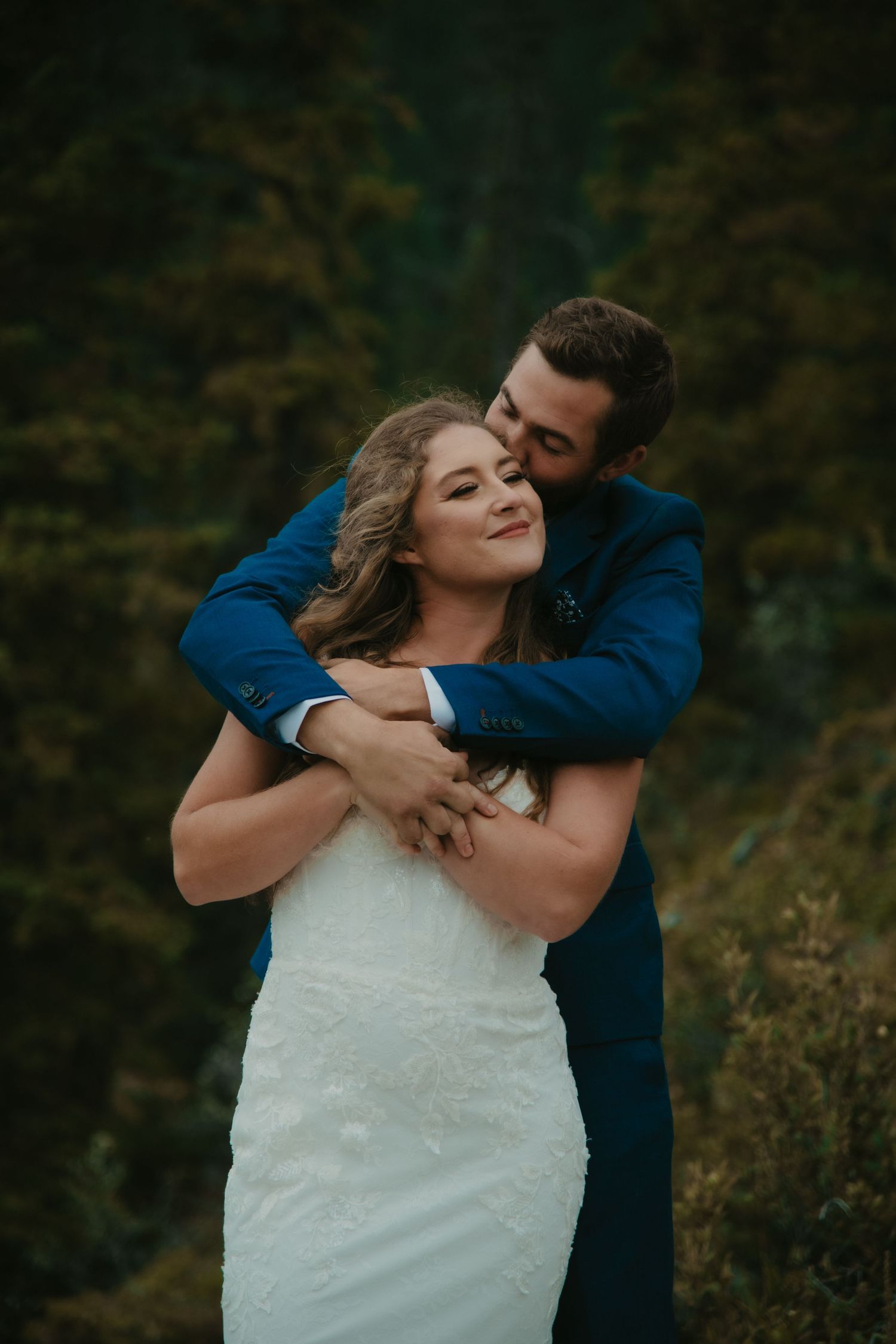 A couple in wedding attire shares an intimate moment against a moody forest backdrop. Calgary Wedding Photographer