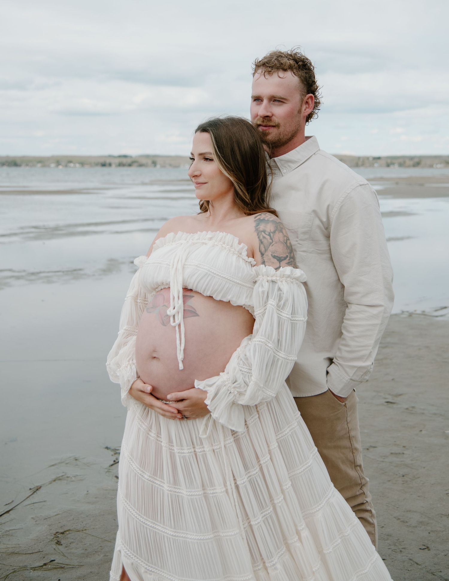Maternity beach photoshoot featuring a couple in light-coloured clothing against a cloudy seascape. Calgary Maternity Photographer