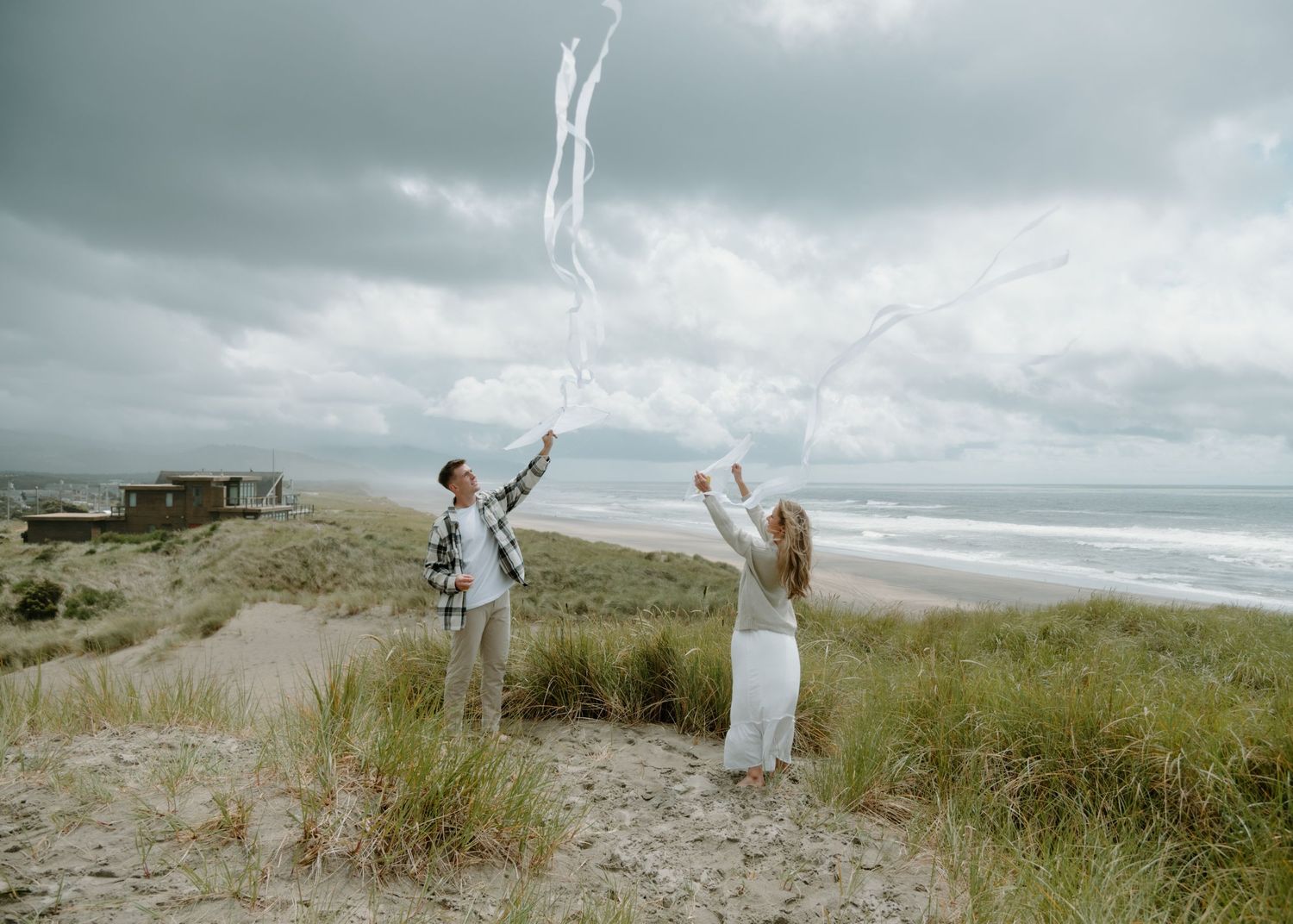 Two people toss flowing white fabric into the air on a dramatic beach clifftop on Oregon Coast. Calgary Lifestyle and Engagement.