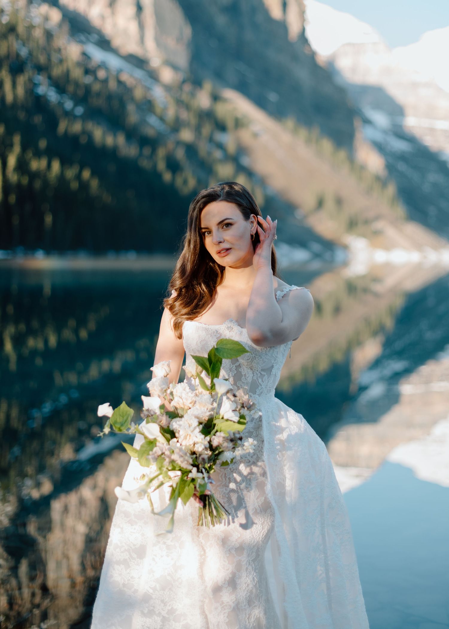Bride holding white bouquet against mountain lake backdrop in dramatic landscape at Lake Louise. Calgary and Banff Wedding Photographer