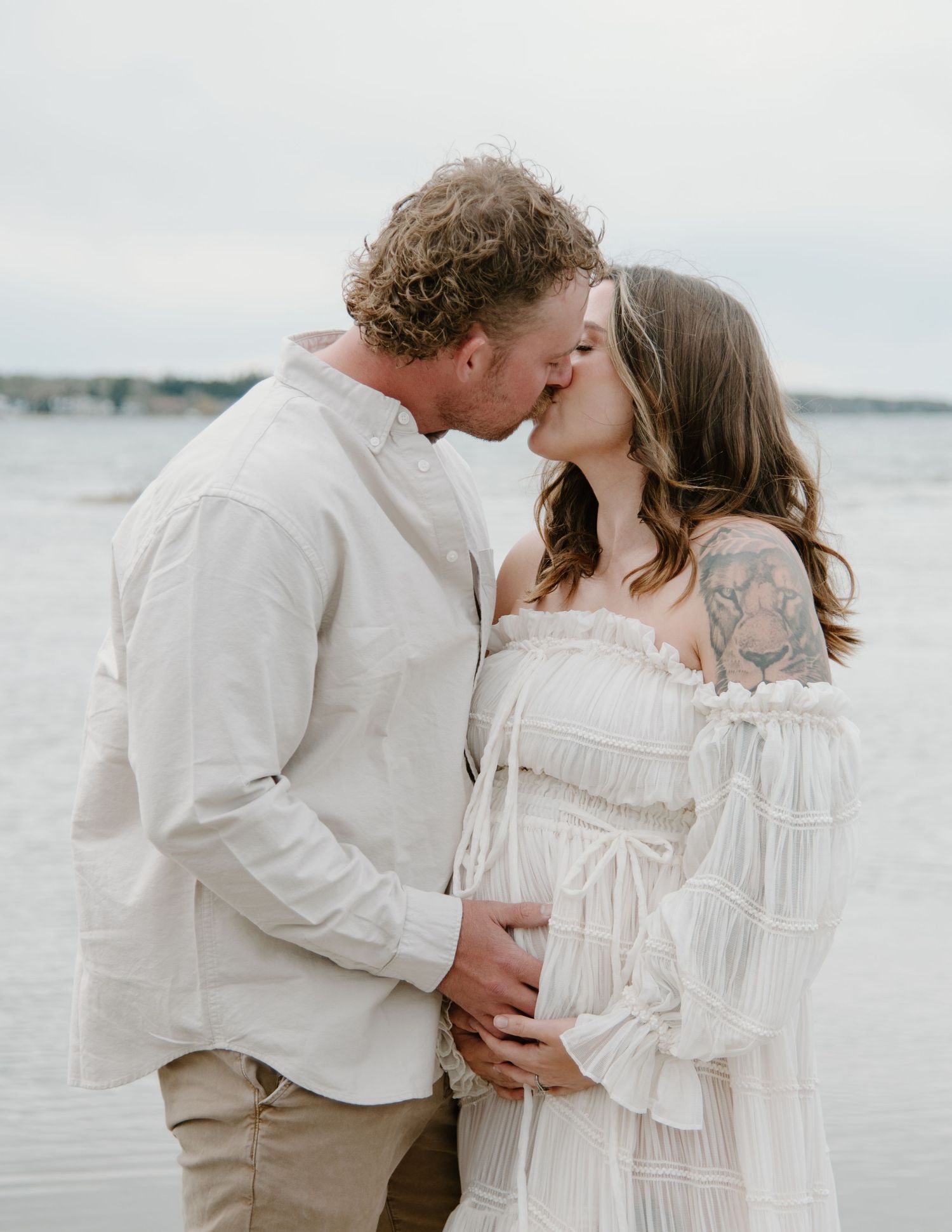 A romantic maternity photoshoot on a beach with a couple in white and beige clothing embracing by the water. Calgary Alberta Maternity Photo