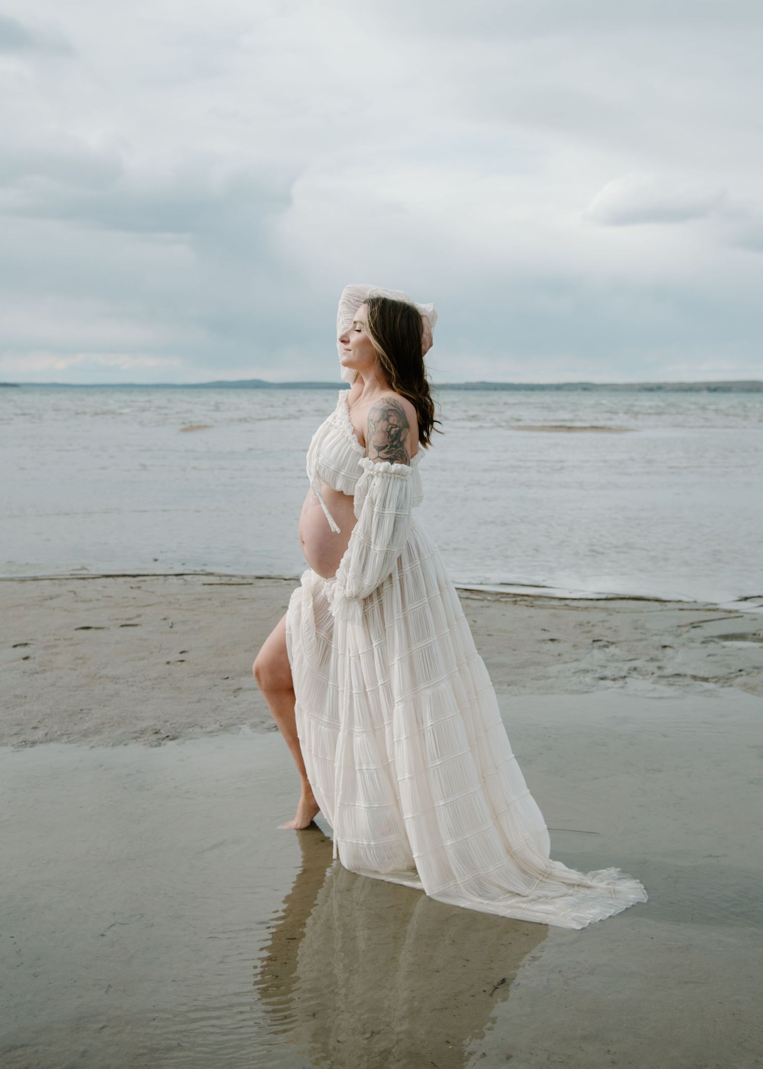 Ethereal maternity photoshoot on a beach with white flowy dress reflecting in wet sand. Calgary Alberta Maternity Photographer.