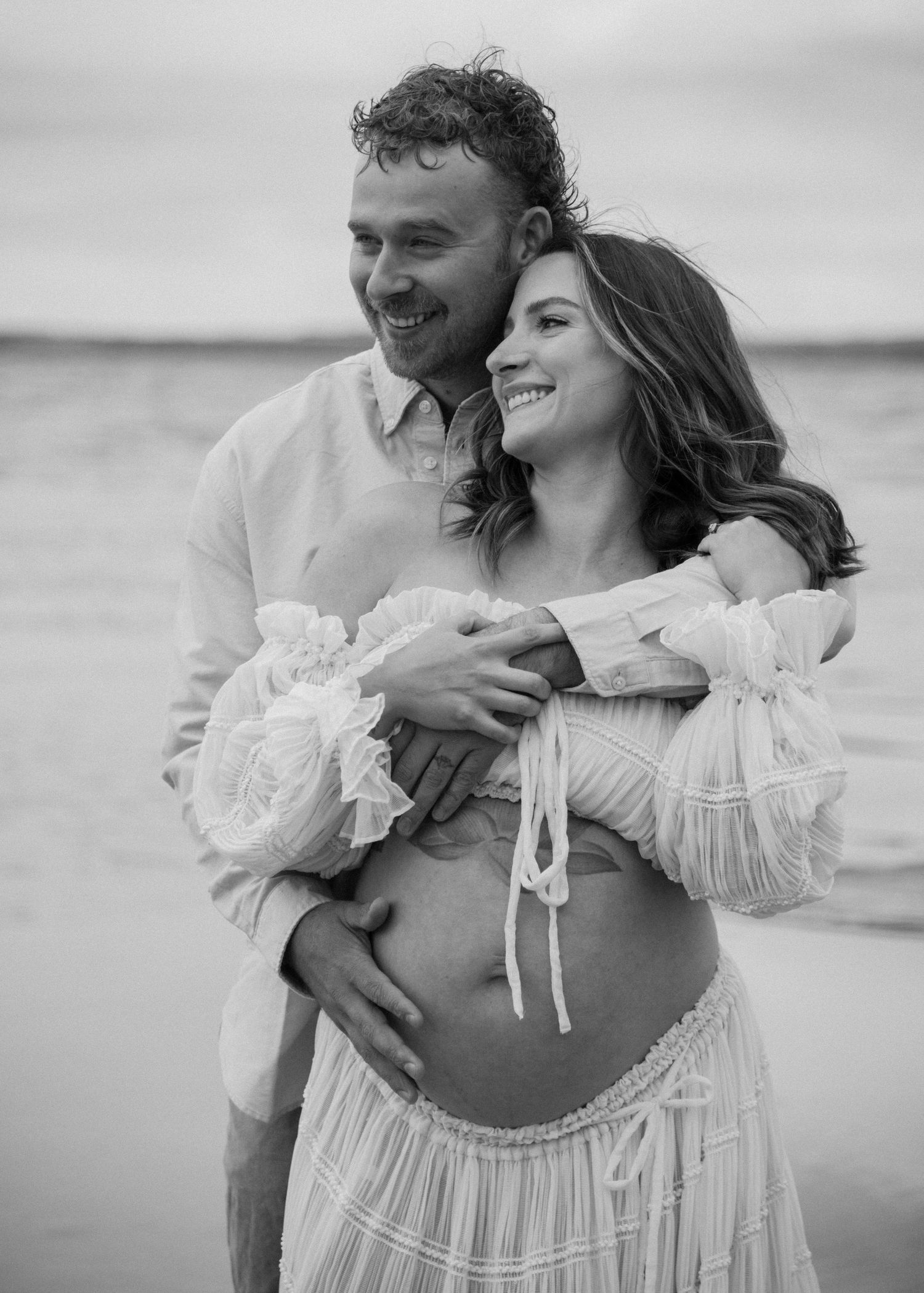 Intimate maternity photoshoot with couple embracing on a beach in white flowing dress. Calgary Alberta Maternity Photographer.