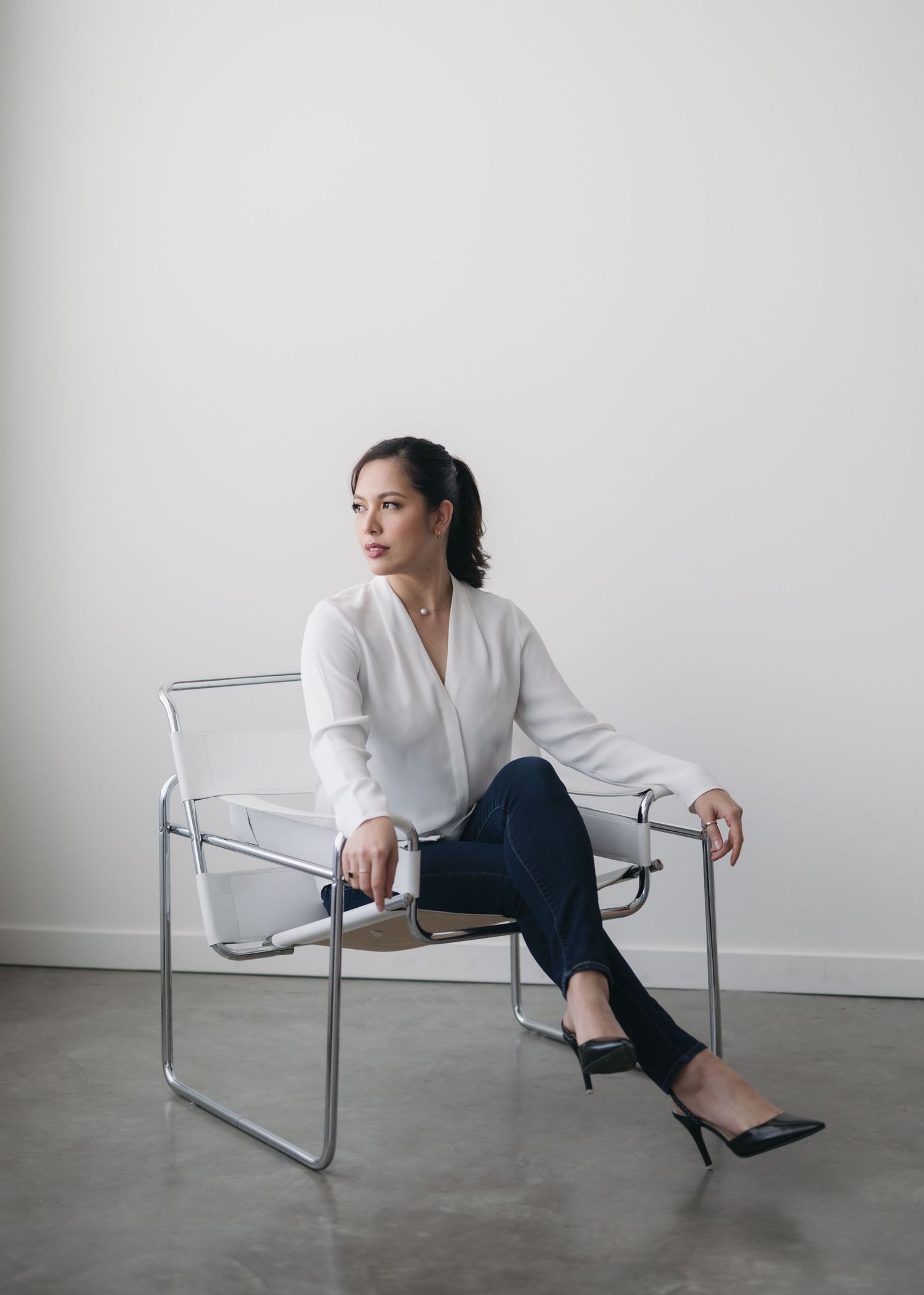 A person in a white blouse and dark pants sits casually on a modern clear acrylic chair against a white wall.