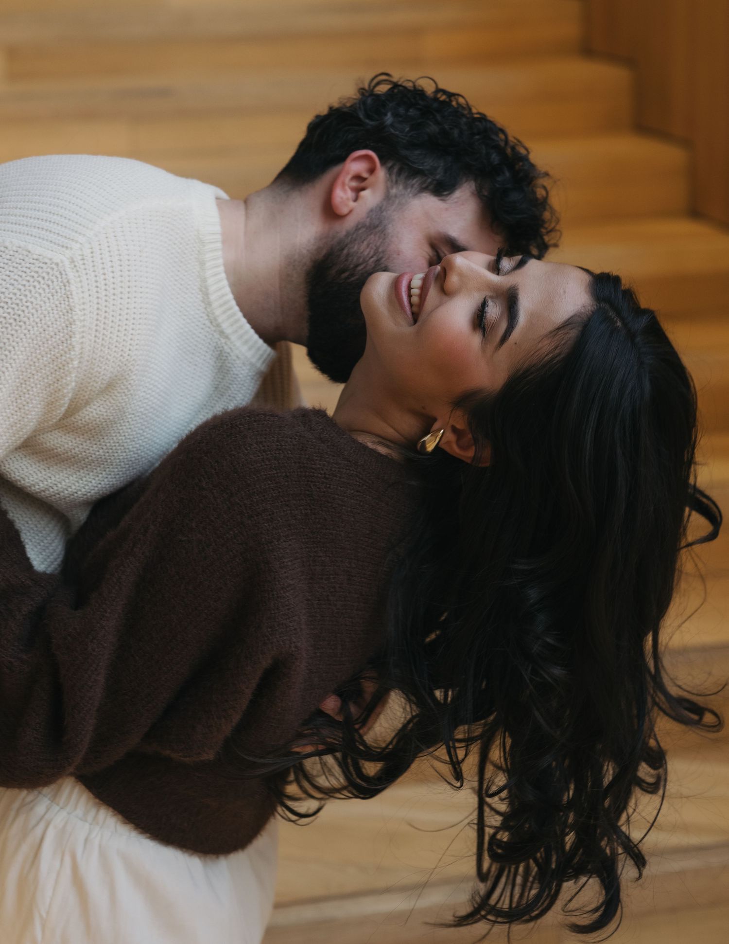A romantic couple in white and brown clothing share an intimate embrace while lying down on a wooden floor.