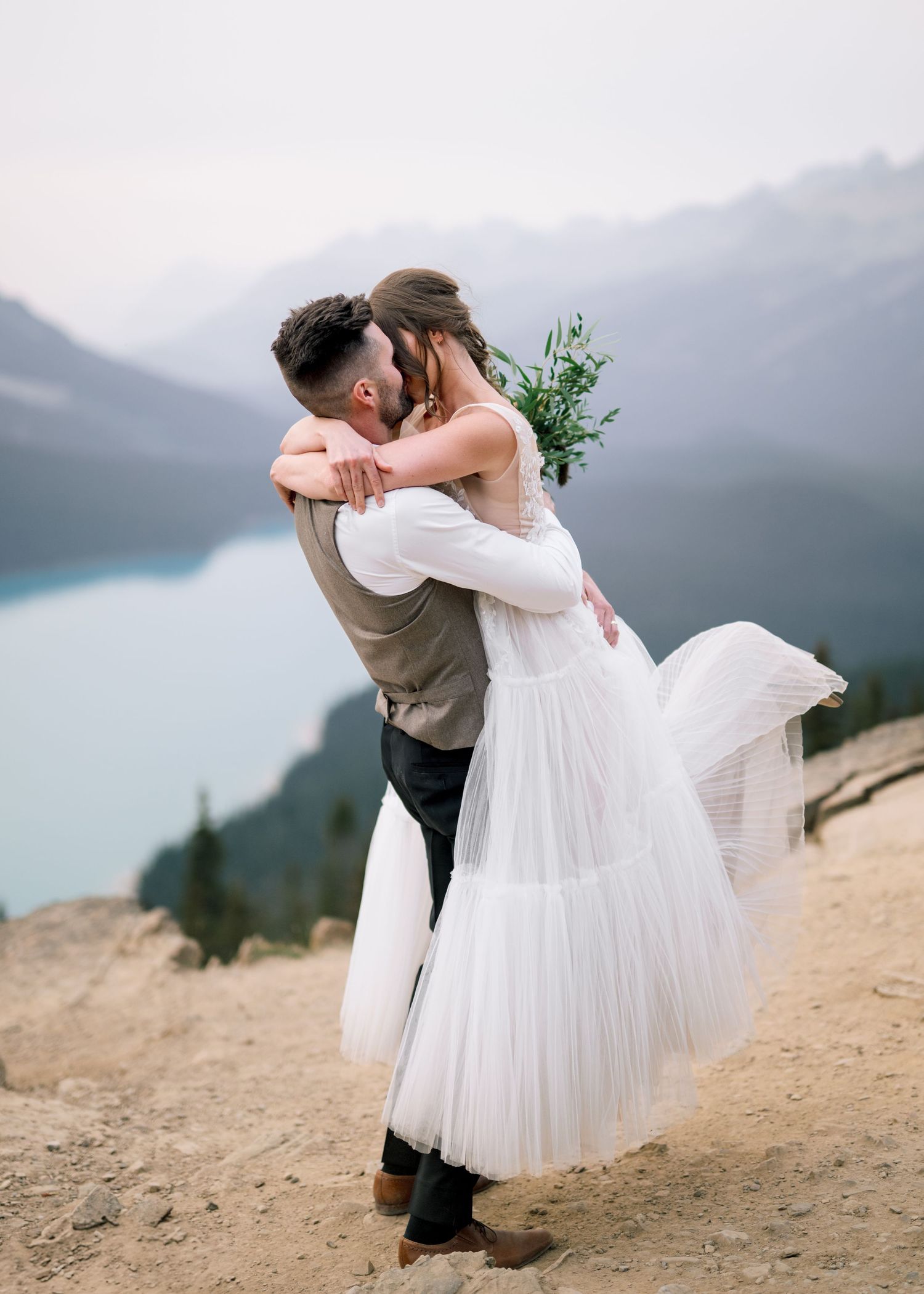 A couple in wedding attire dance and embrace on a scenic mountain cliff overlooking a serene lake.