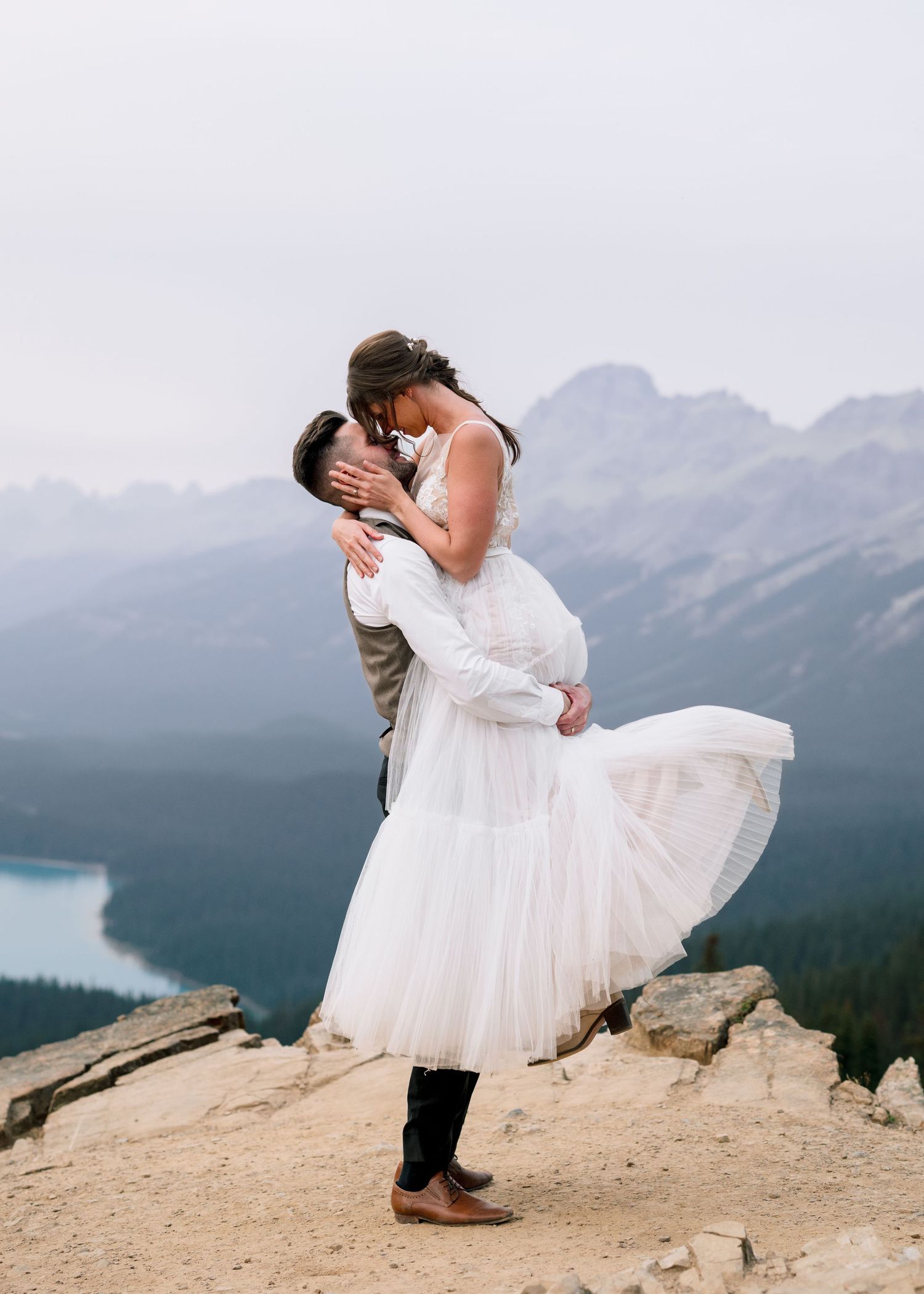 Newlywed couple sharing a romantic kiss on a mountain overlook with a stunning lake and mountain vista in the background.
