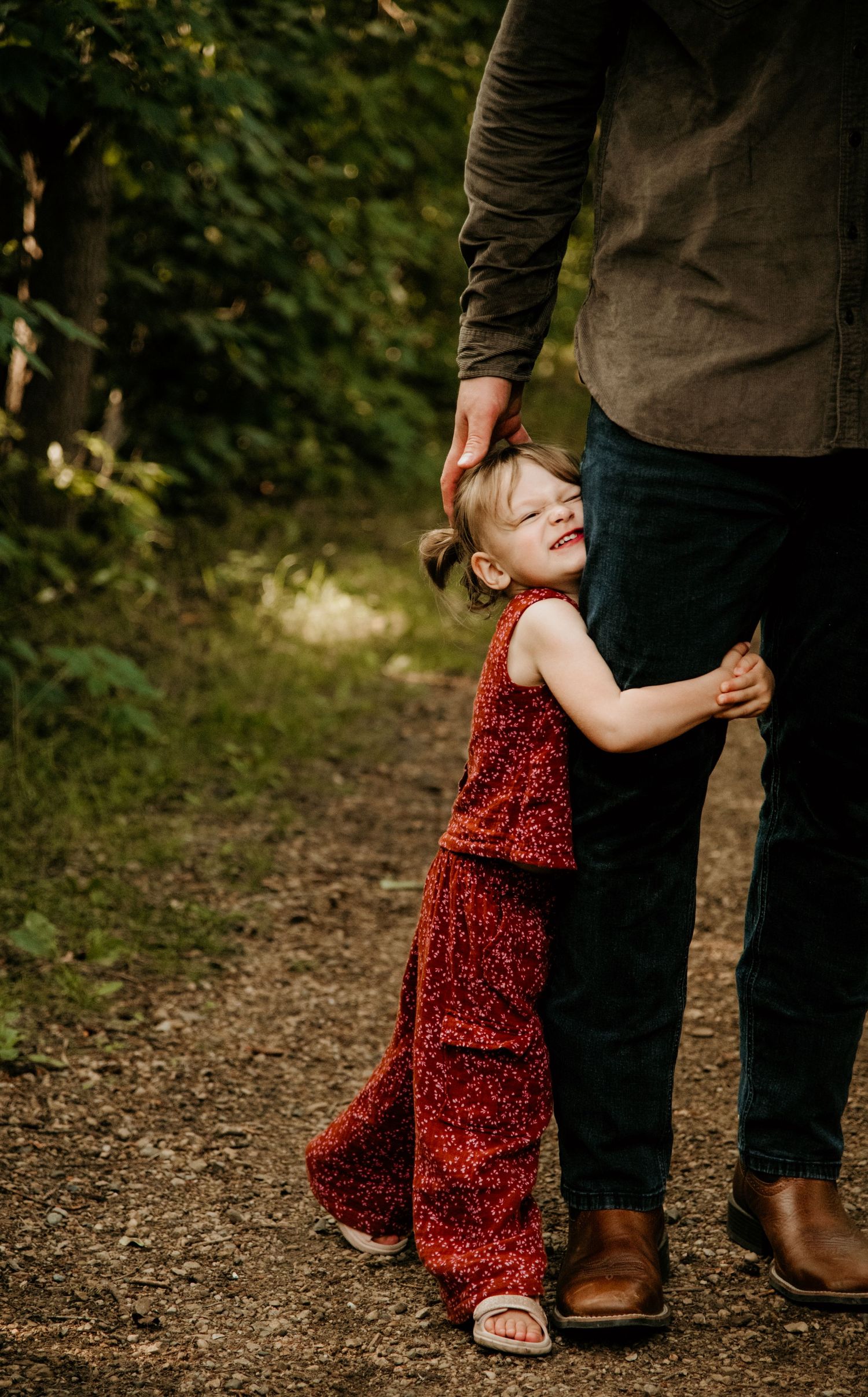 A child in a sparkly red outfit hugs an adult's legs on a forest path. Calgary Alberta Family Photographer.