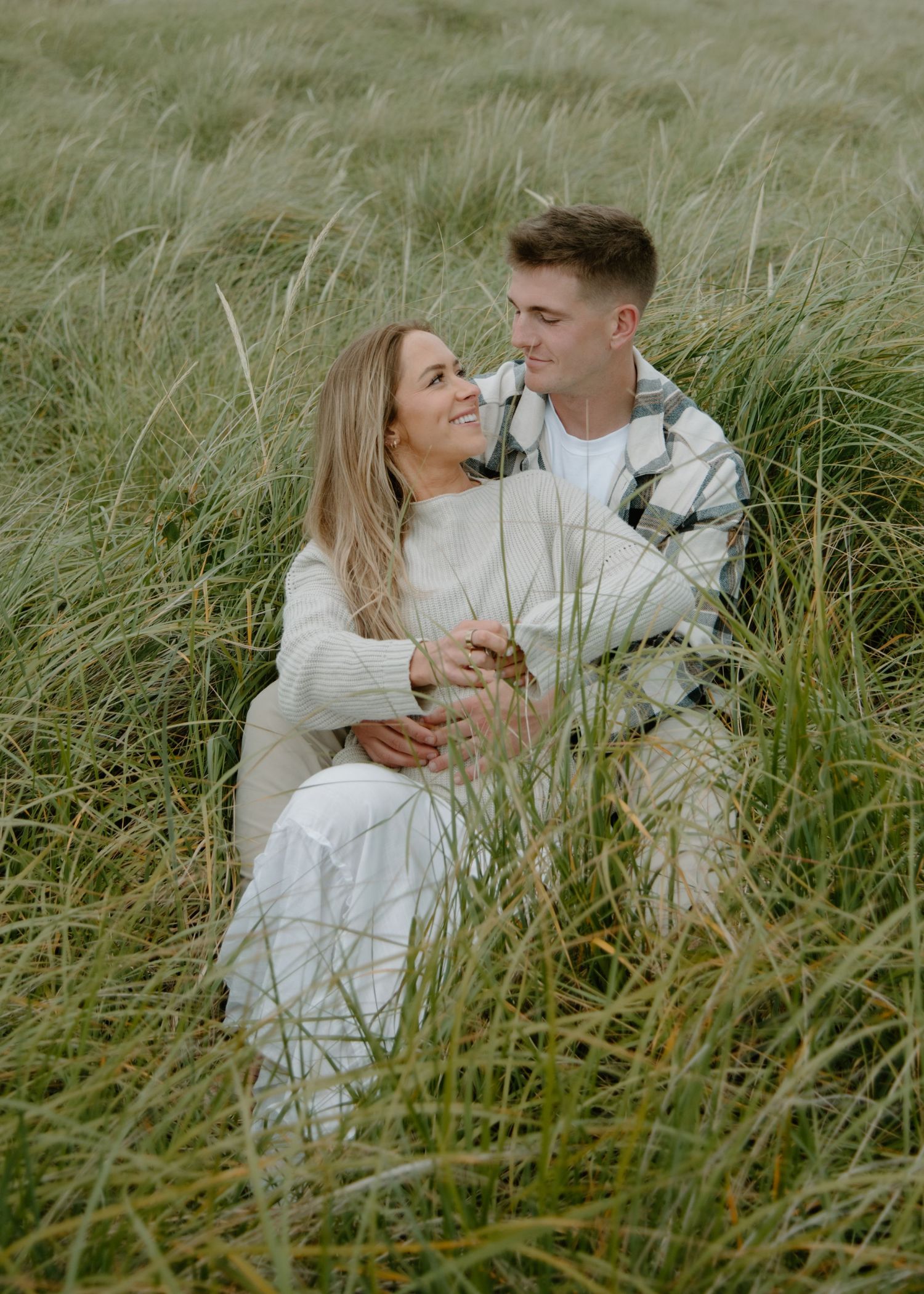 Couple in matching white outfits share loving embrace among coastal grass on Oregon Coast. Calgary Lifestyle and Engagement Photographer.