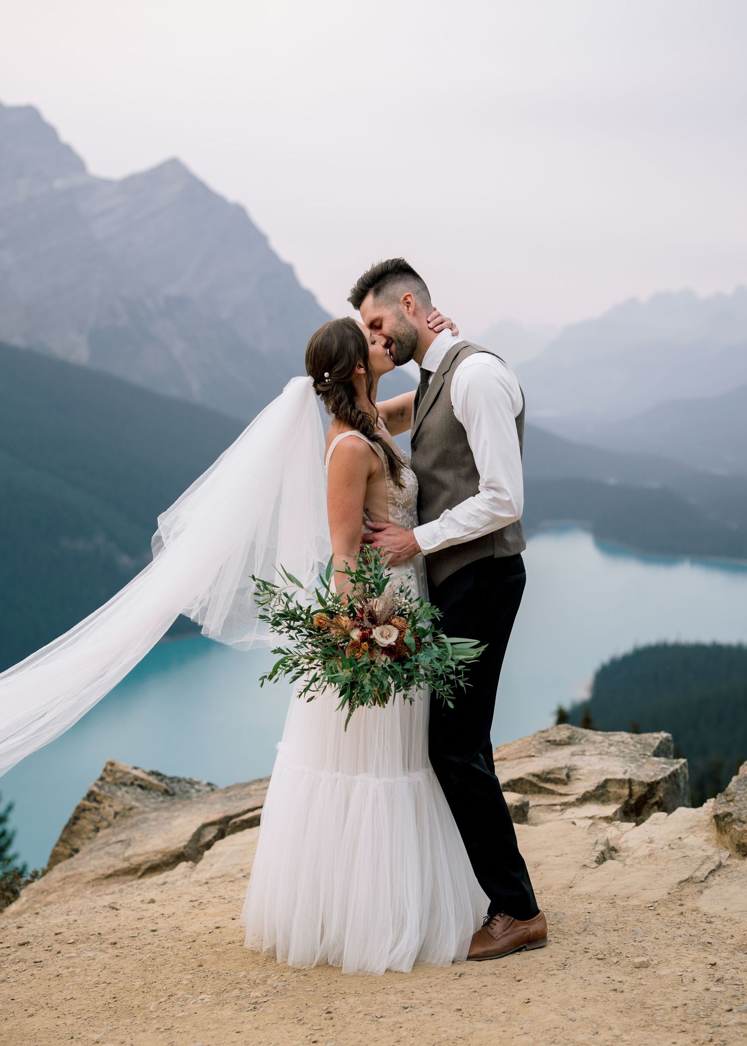 A romantic wedding portrait shows a couple kissing on a mountain overlook with a turquoise lake and peaks in the background.