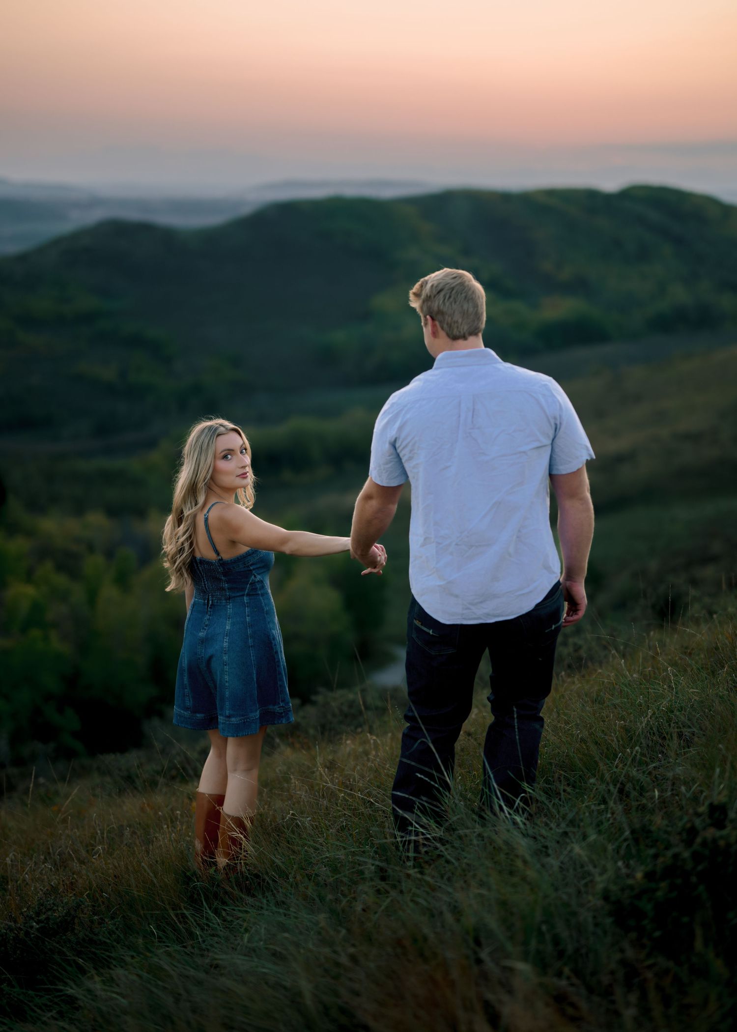 Two people walk through a scenic hillside field at sunset, creating a silhouetted scene against the dusky sky.