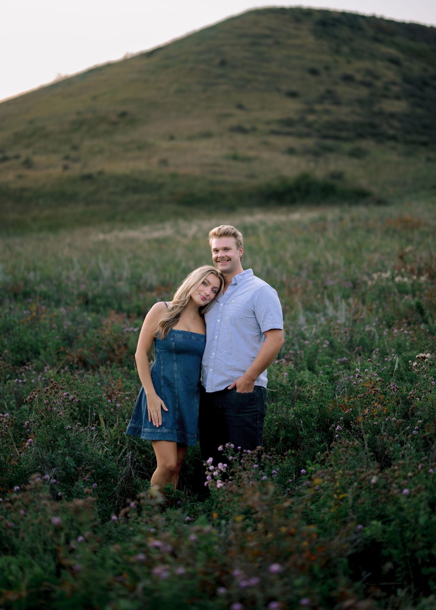 A couple shares a tender moment while sitting together in a wildflower meadow with rolling hills in the background at sunset.