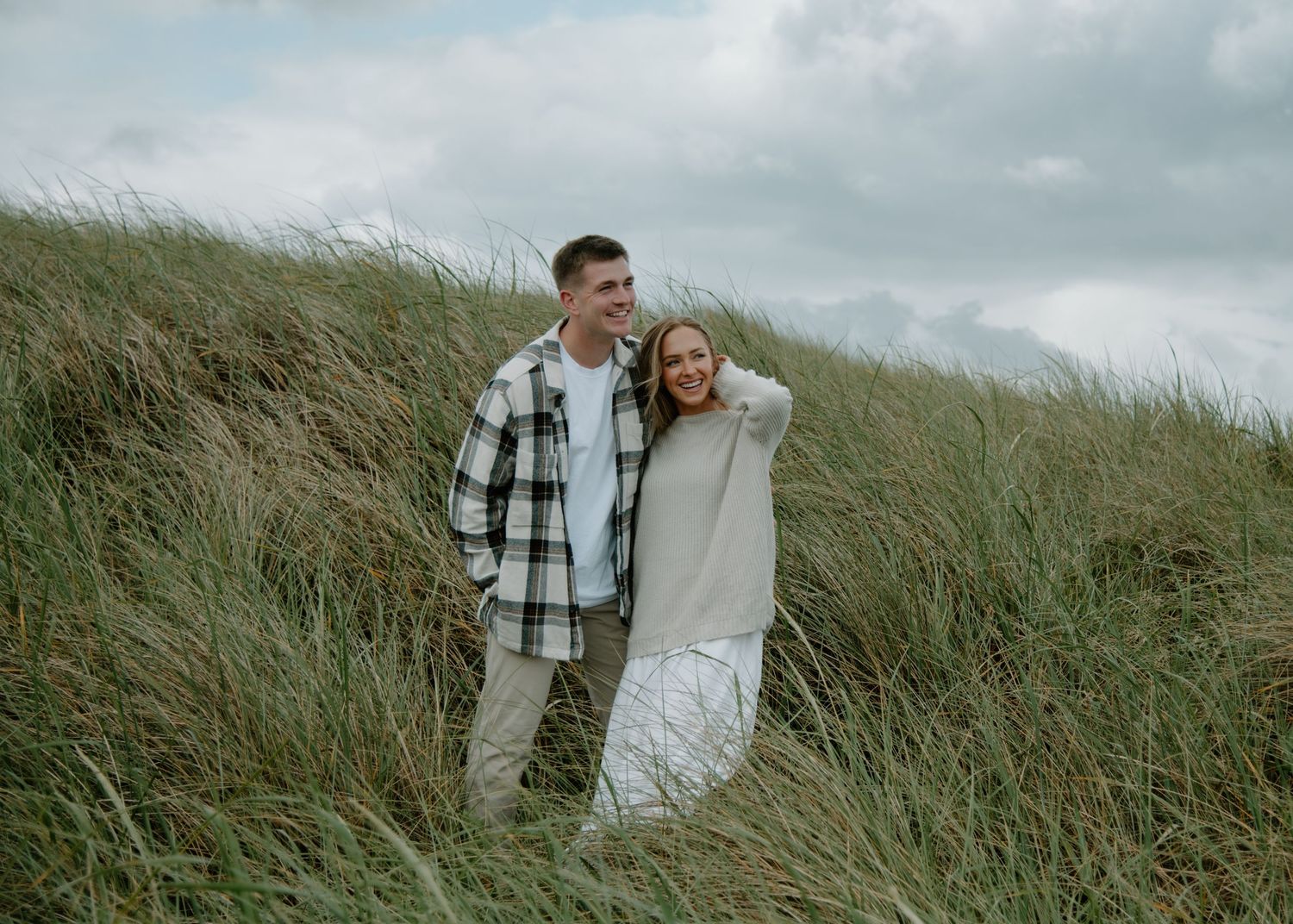 A couple stands together smiling among windswept beach grass on a cloudy day on Oregon Coast. Calgary Lifestyle and Engagement Photographer.