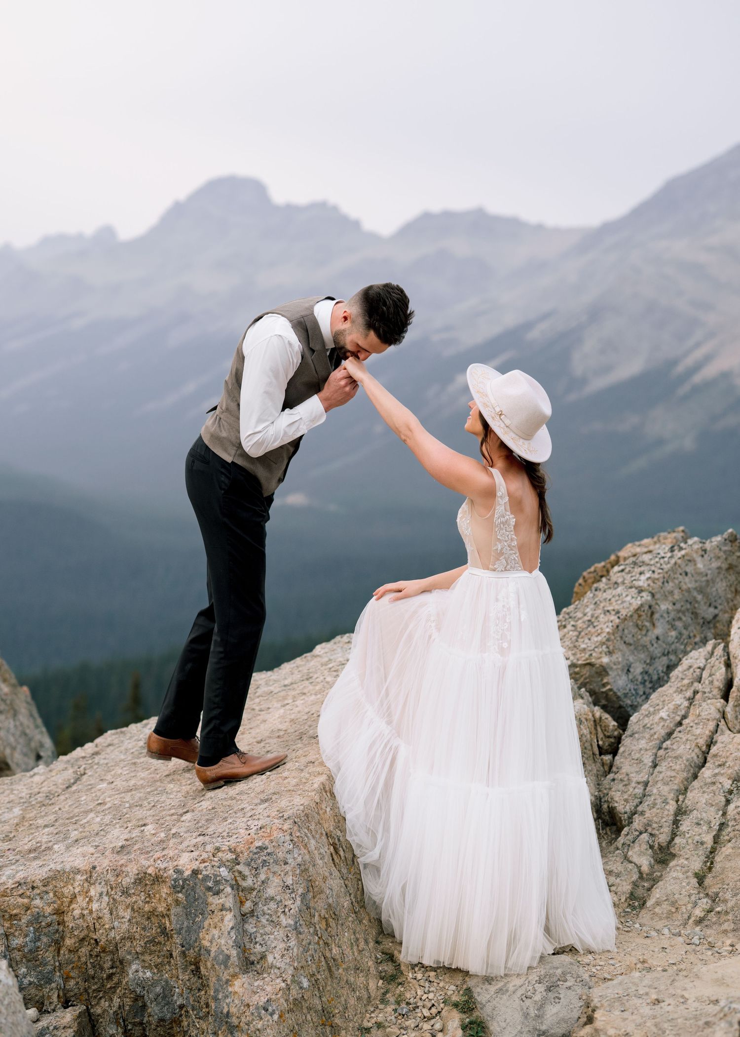 A couple in wedding attire embraces on a mountain peak with misty mountain ranges stretching into the distance.