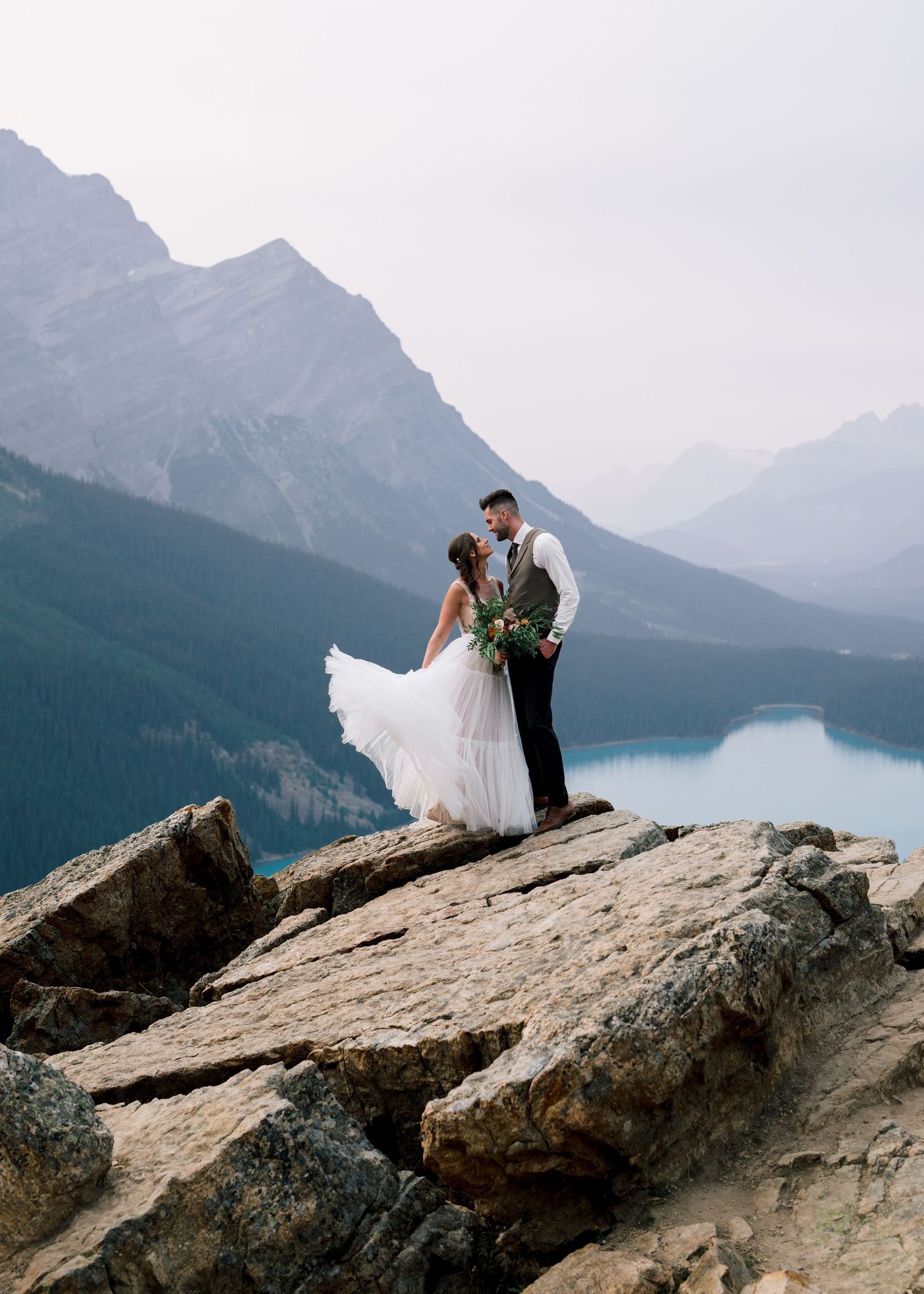 A couple in wedding attire stands on a rocky cliff overlooking a stunning mountain lake in Banff National Park at sunset.