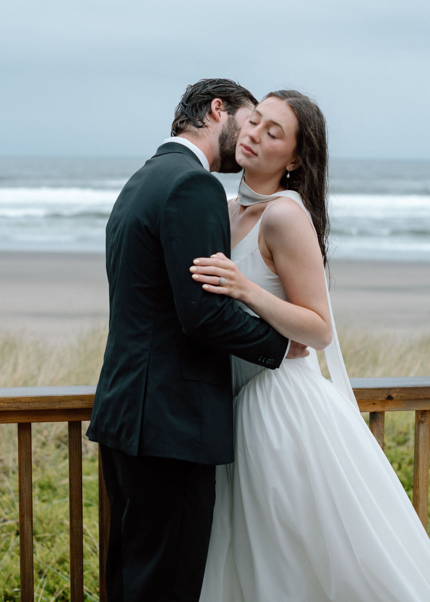 Couple in wedding attire share a romantic kiss on the beach boardwalk while gray ocean waves roll in behind them.