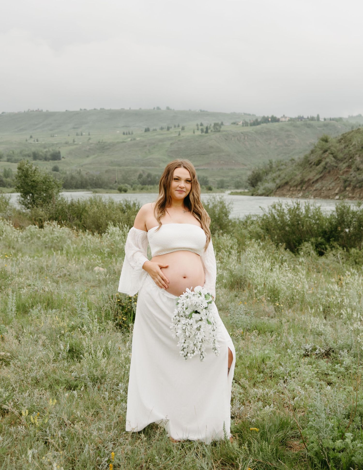 A pregnant woman in a flowing white dress stands in a green meadow with mountains in the background.