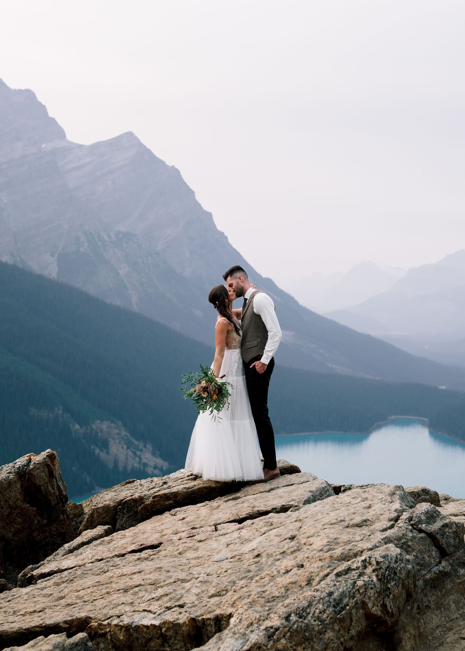 A couple in wedding attire share an intimate moment on a rocky cliff overlooking a misty mountain lake.