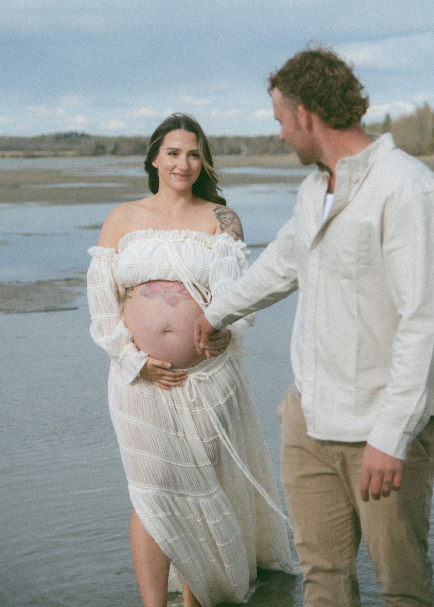 A romantic maternity photoshoot on a beach with a couple in white clothing embracing and showing the pregnant belly. Calgary Photographer