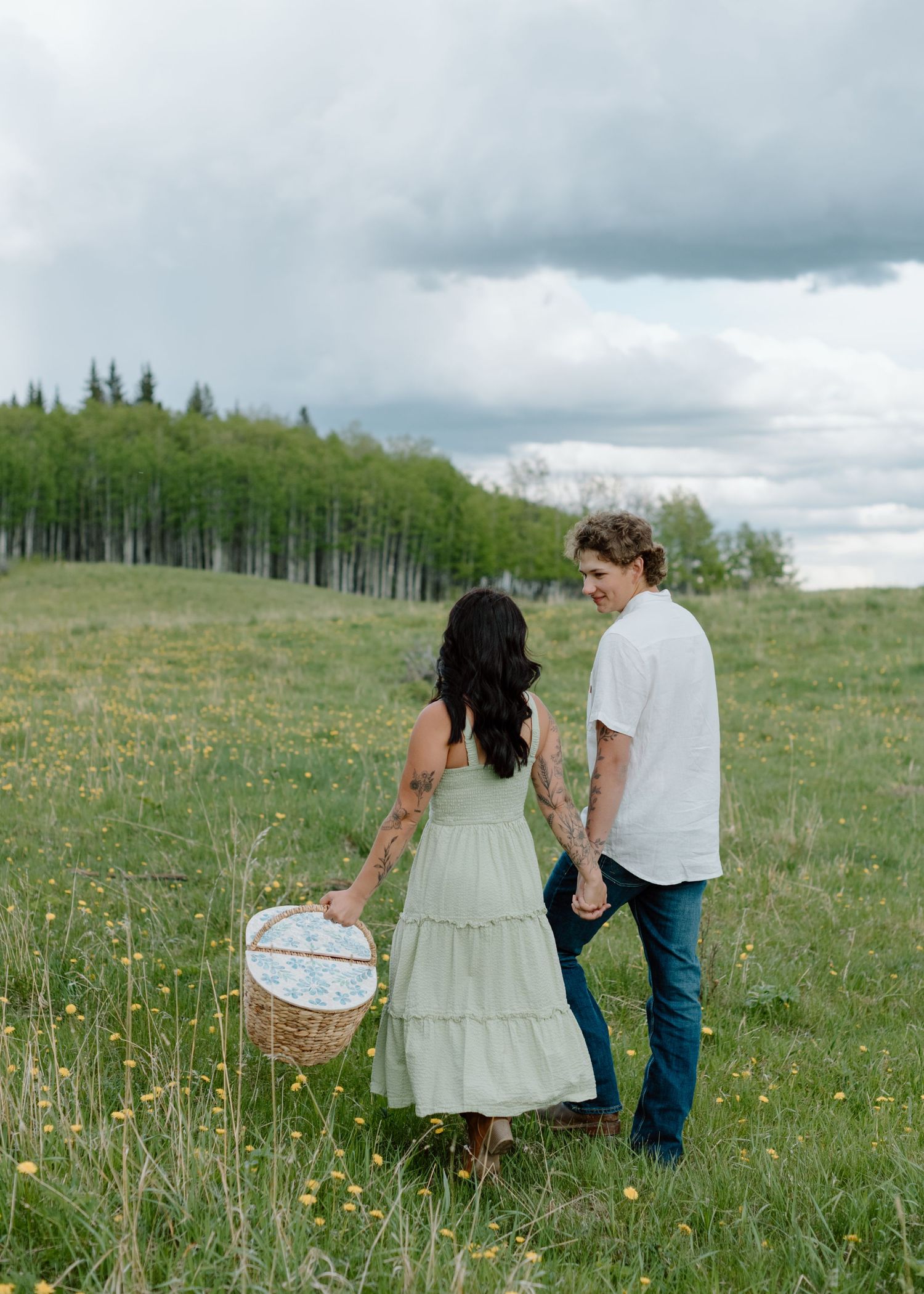 Two people walking through a wildflower meadow with baskets. Calgary Alberta Engagement Photographer.
