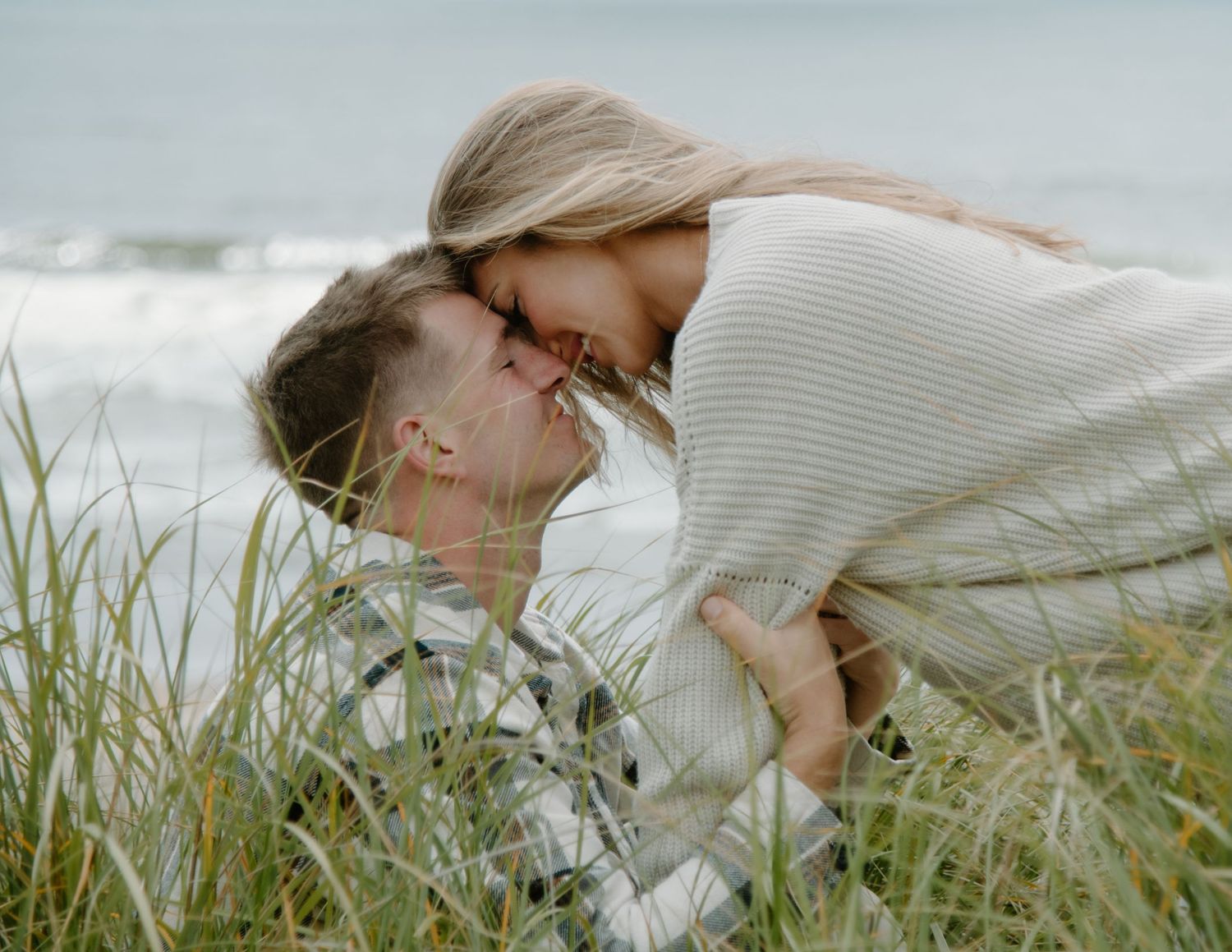 Romantic embrace between couple nestled in beachside grass on Oregon Coast. Calgary Lifestyle and Engagement Photographer.