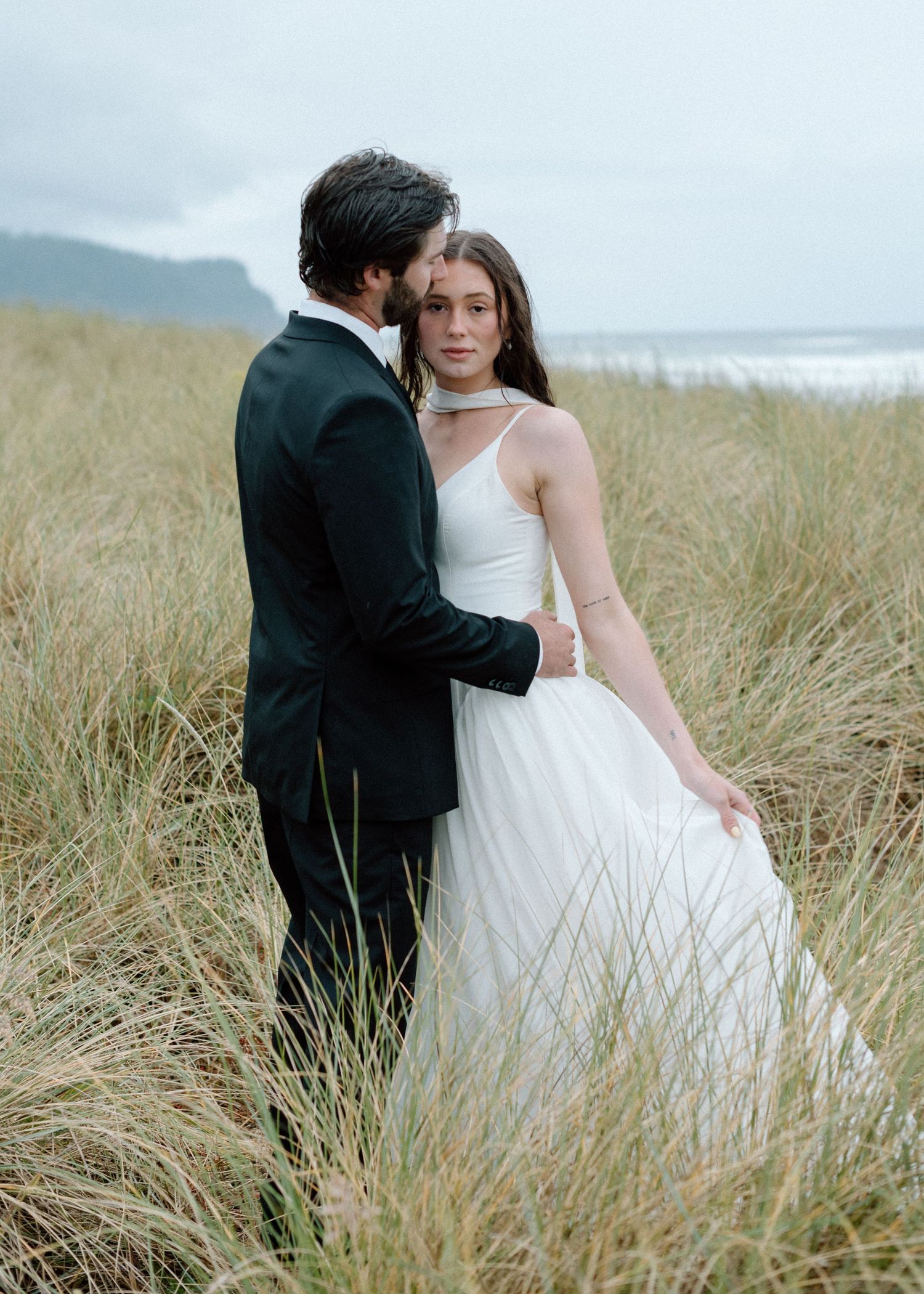 A romantic wedding couple embraces in tall beach grass along the shoreline on their special day.