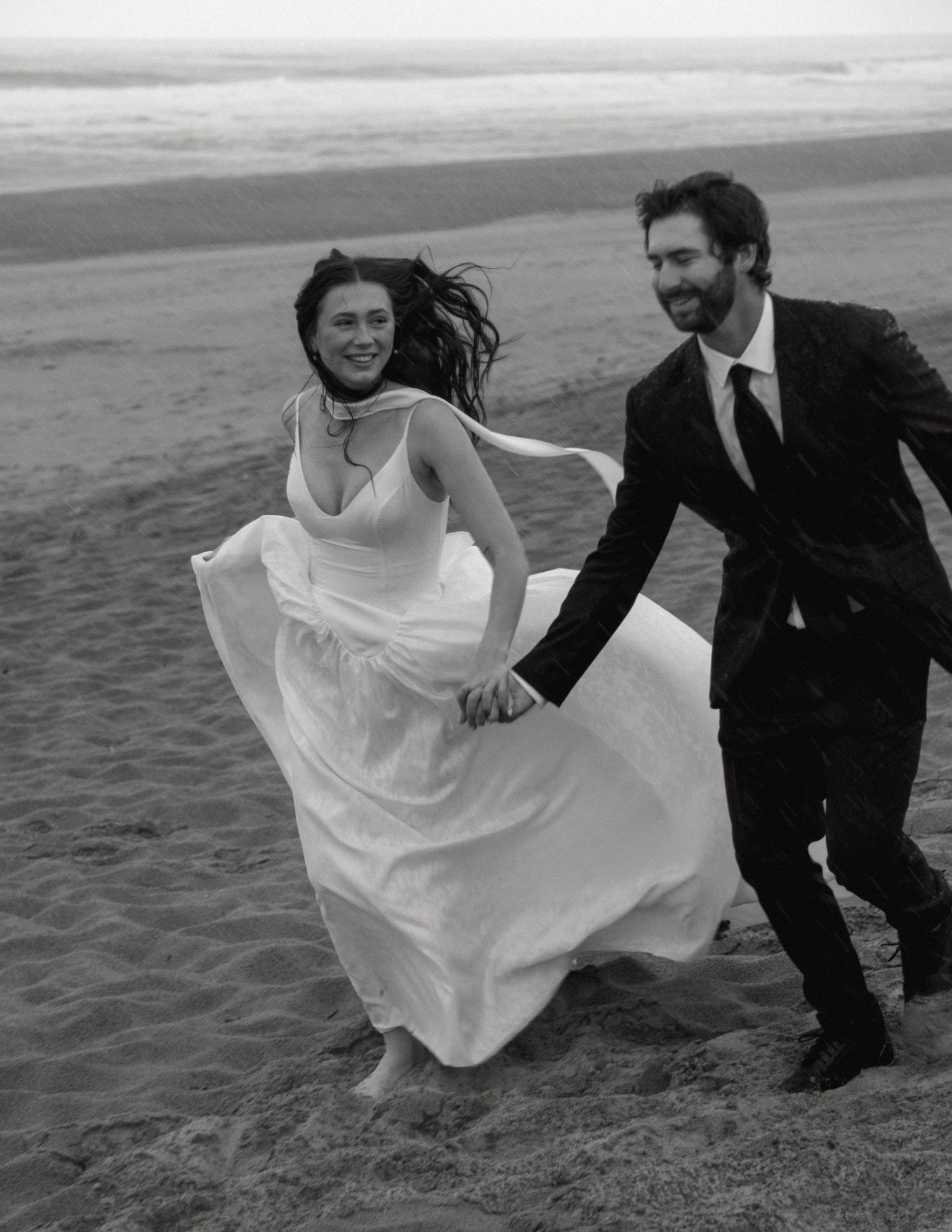 A couple in wedding attire running joyfully on a beach, with the bride's white dress flowing in the wind.