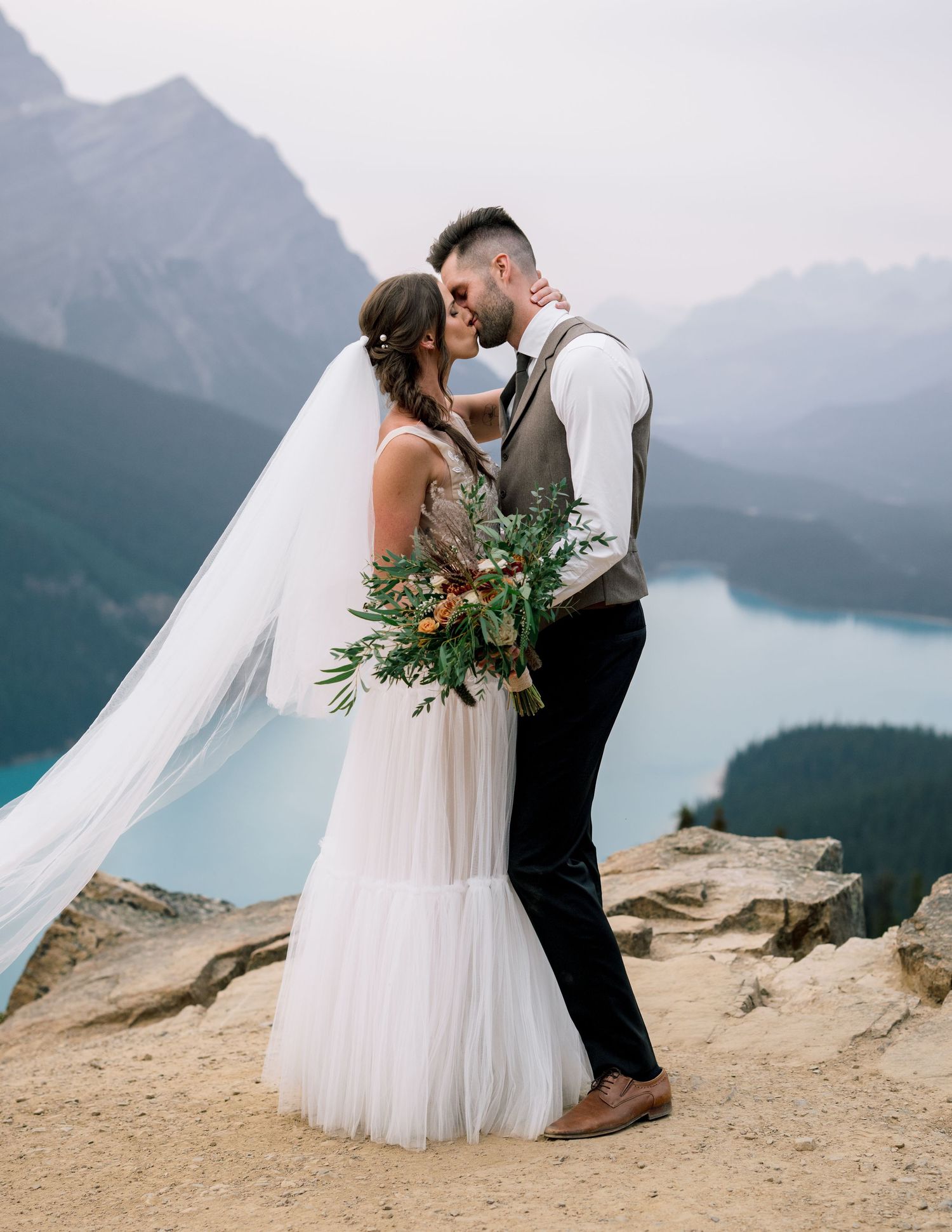 A romantic wedding ceremony on a mountain cliff overlooking a misty turquoise lake and mountain ranges.